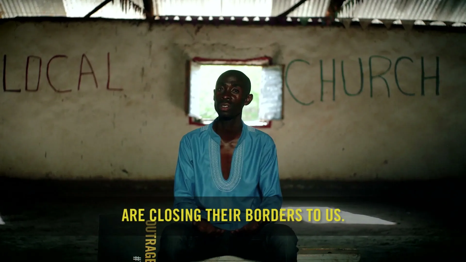 A young Black man in a blue shirt sits in a dimly lit, rustic room with "LOCAL CHURCH" painted on the wall, speaking to the camera.