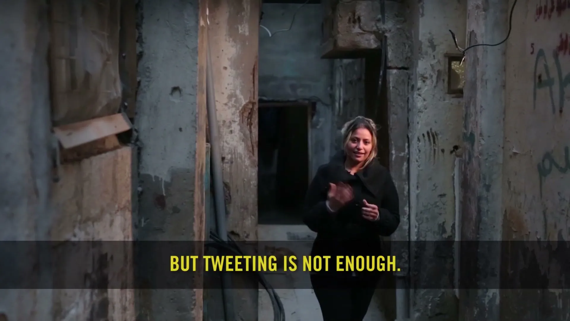 A woman in her 30s, wearing a black coat, stands in a narrow, dilapidated alleyway, speaking directly to the camera.