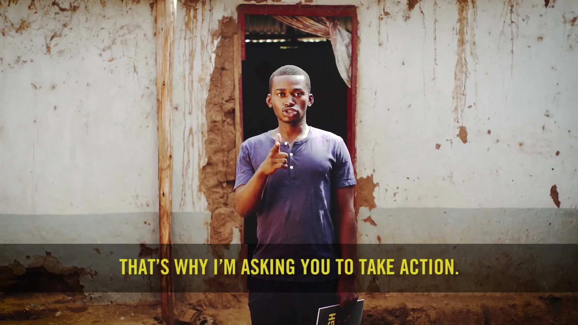 A young Black man in a blue t-shirt stands in front of a dilapidated building, speaking directly to the camera, with text on screen.