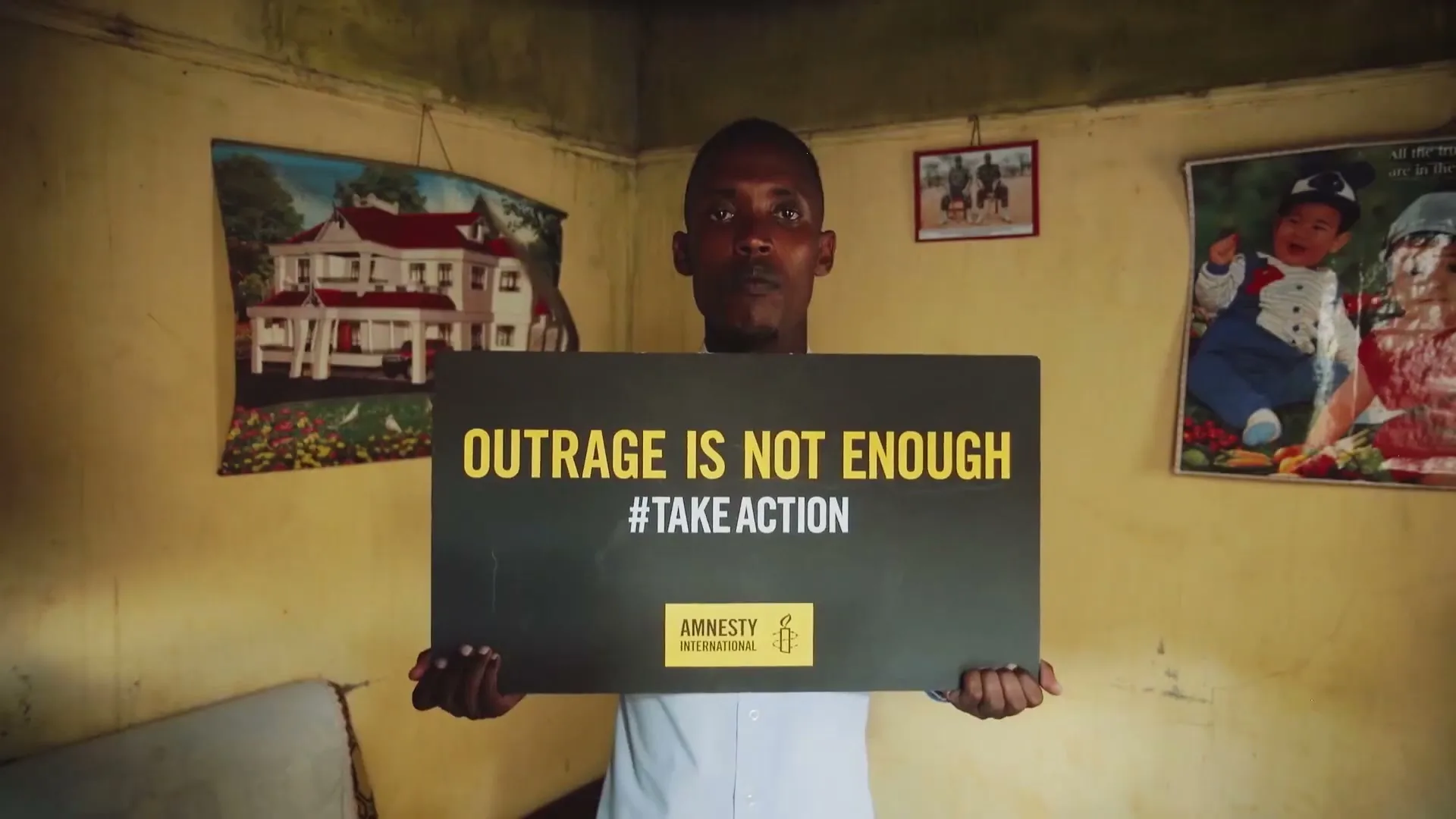 A man in his 20s holds a sign that reads "OUTRAGE IS NOT ENOUGH #TAKEACTION" and "AMNESTY INTERNATIONAL" in a dimly lit room.