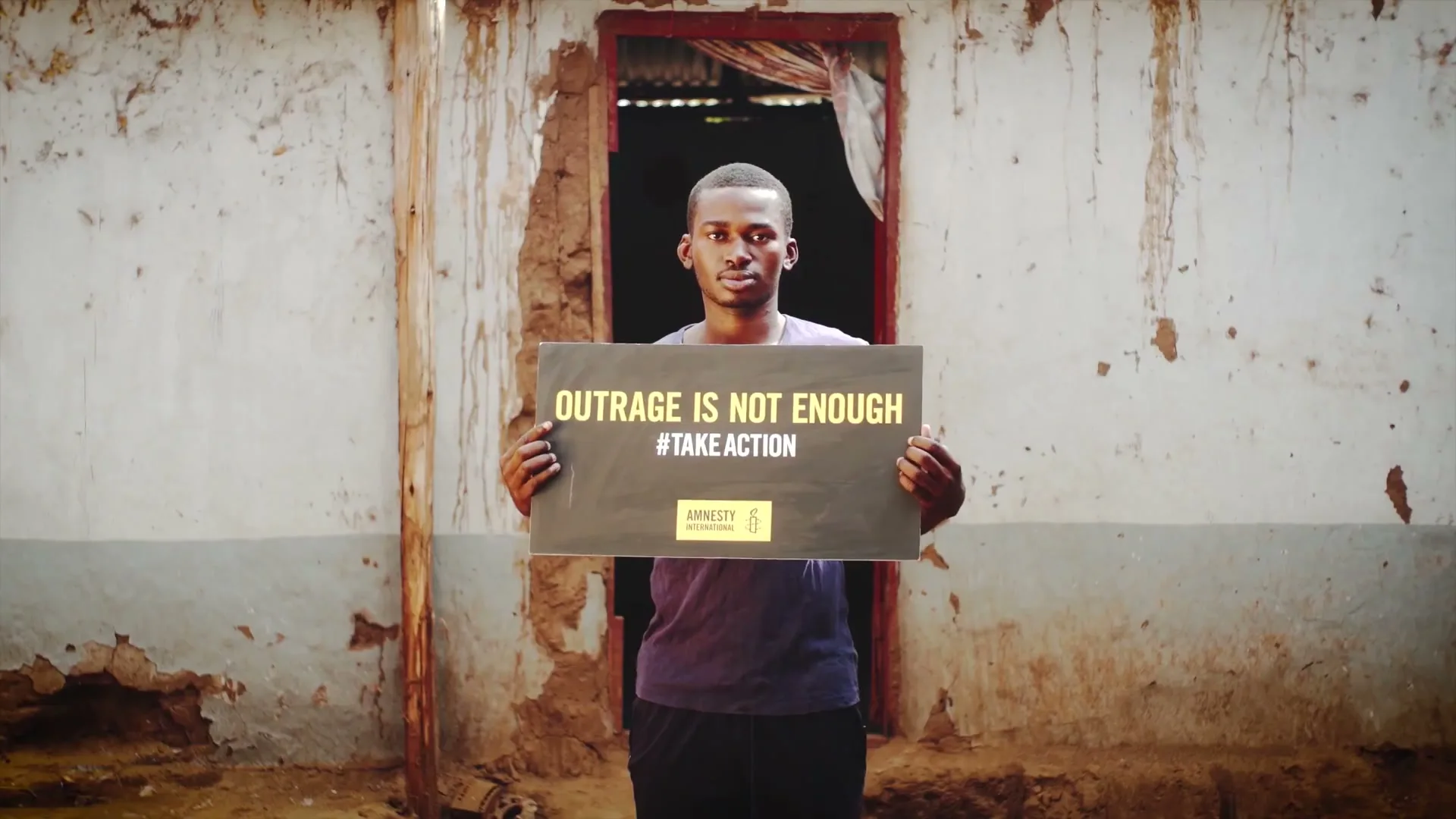 A young man holds a sign that reads "OUTRAGE IS NOT ENOUGH #TAKEACTION" in front of a dilapidated building.