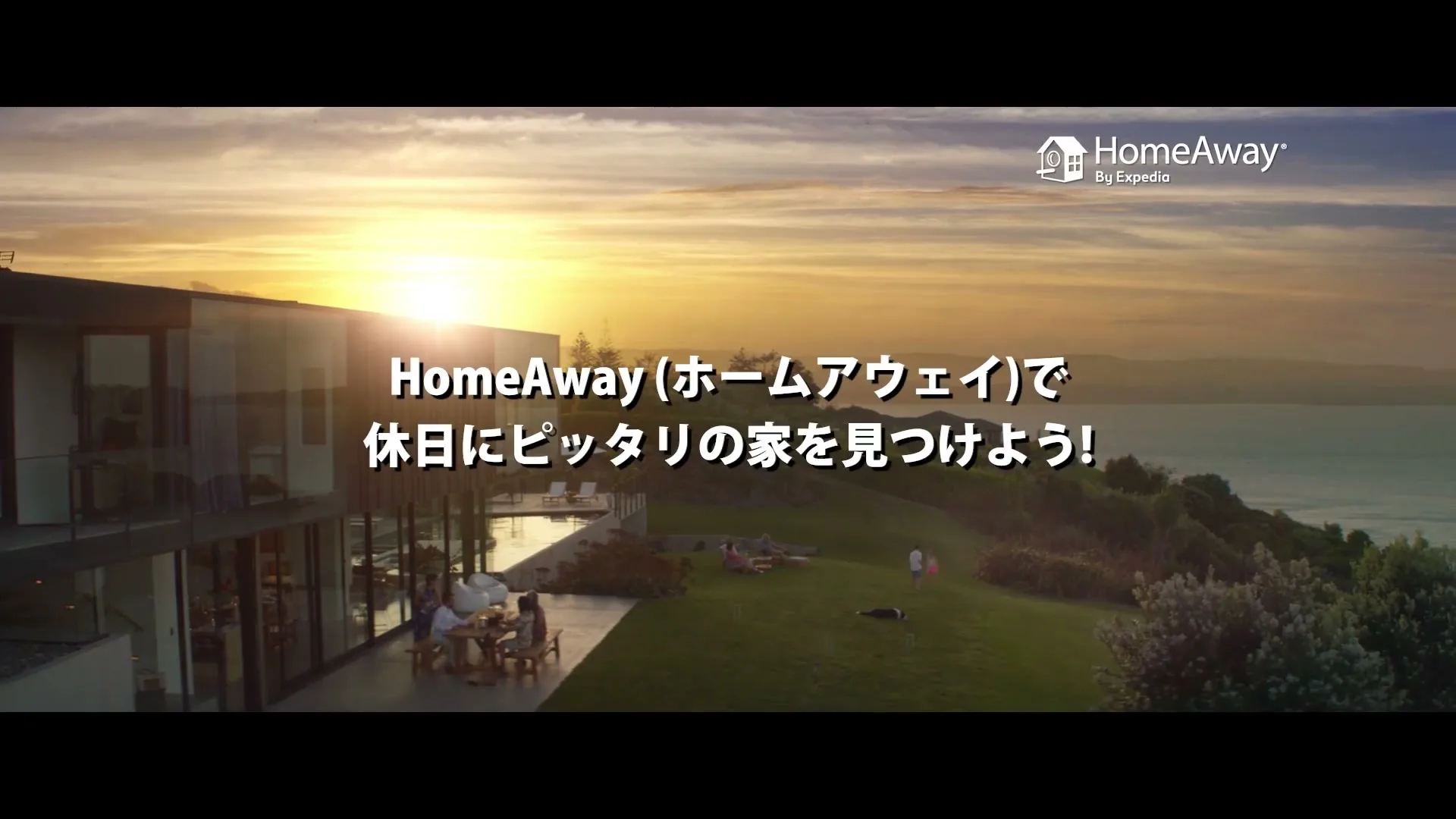 A modern glass house with a patio overlooks the ocean at sunset, with people relaxing on the lawn and patio.
