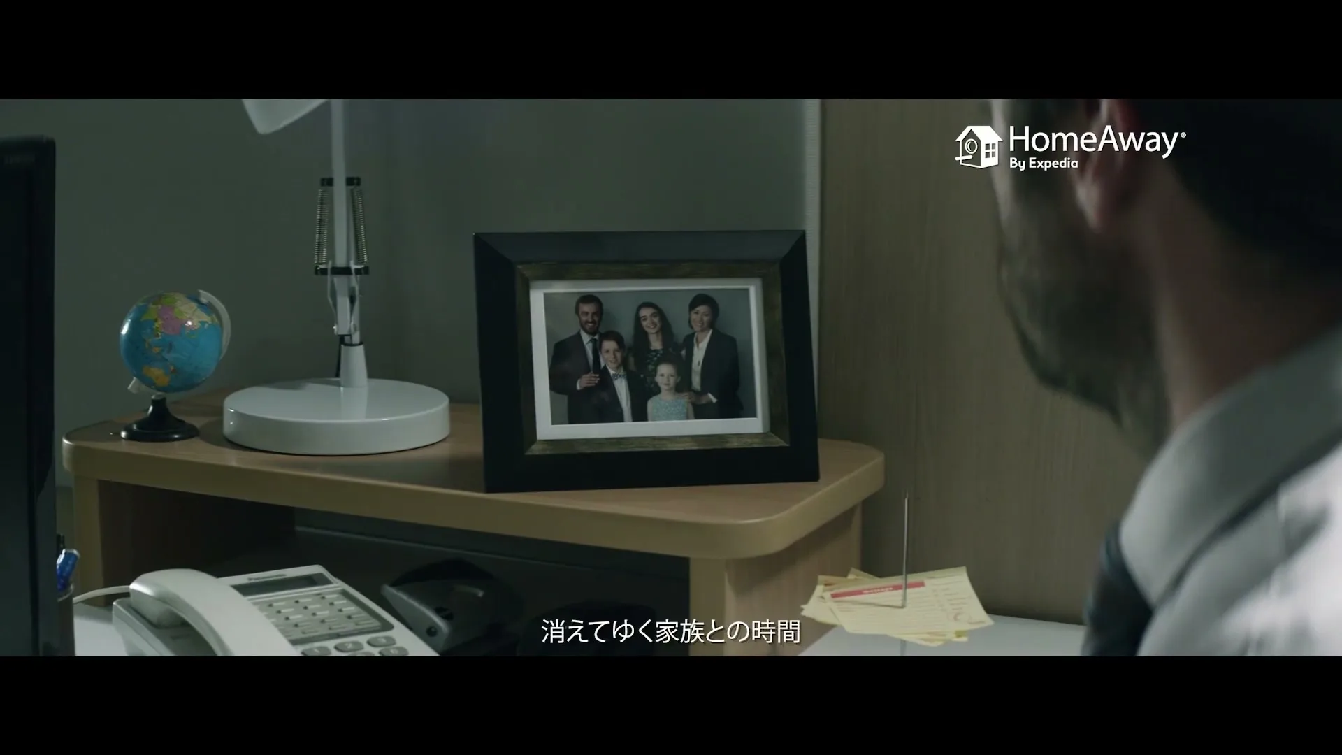 A man looks at a framed family photo on his office desk, with a lamp and a globe nearby, in a dimly lit office.