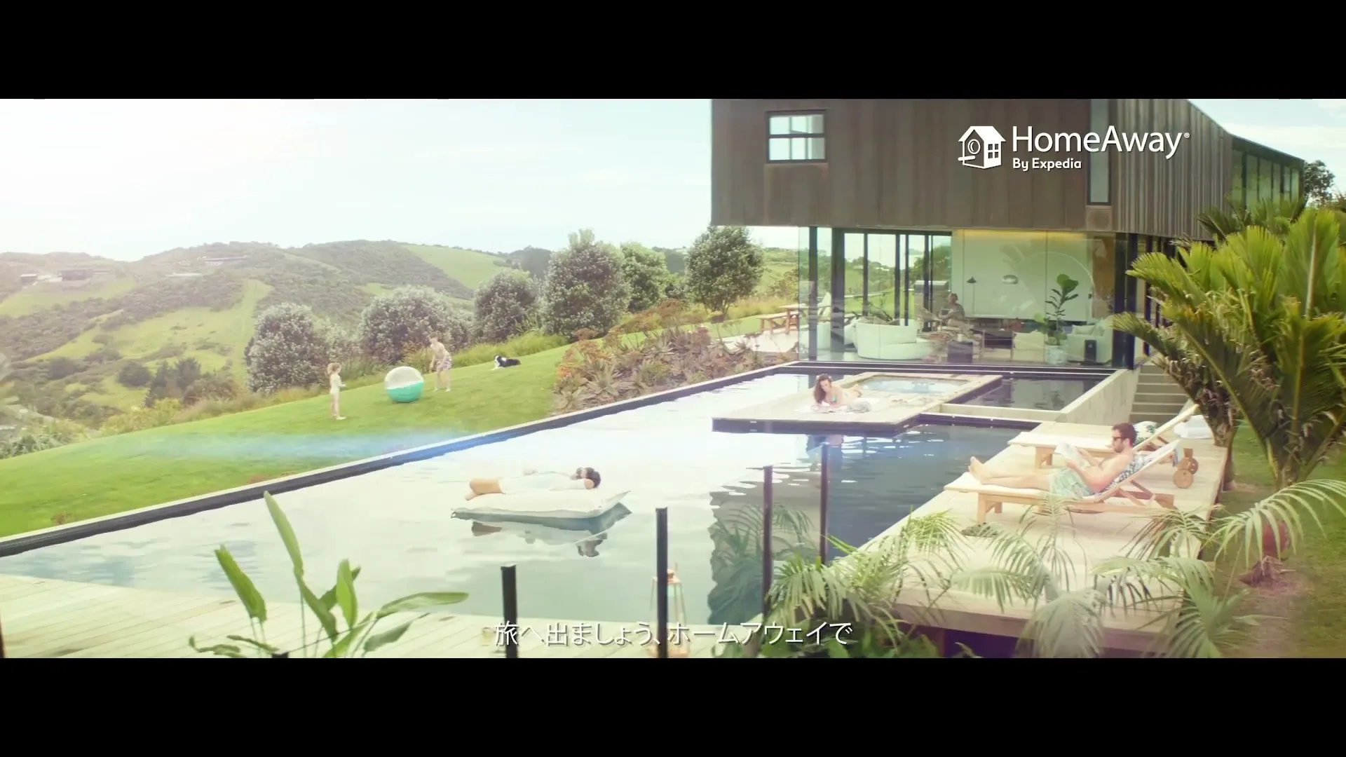 A wide shot shows a modern house with a large pool, where people relax and play on a sunny day.
