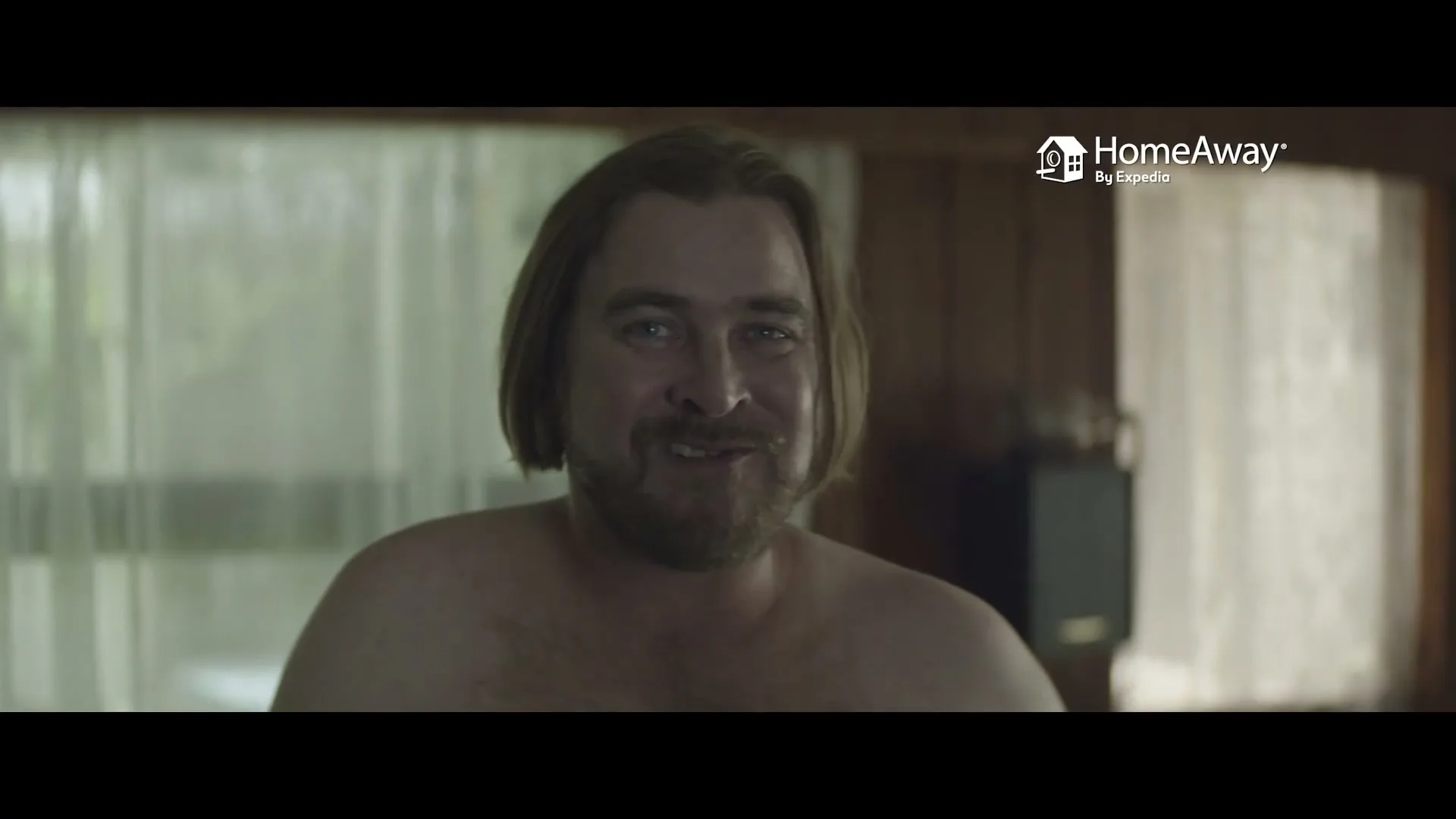 A shirtless man with long hair and a beard smiles at the camera in a dimly lit room with a window in the background.