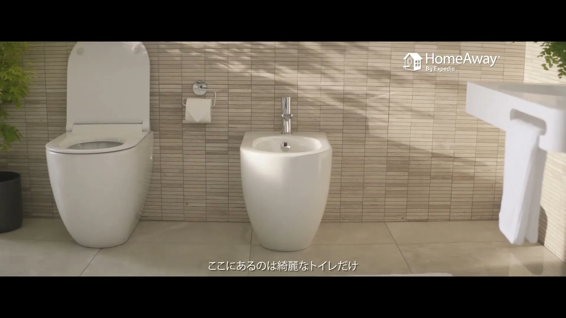 A clean, modern bathroom features a white toilet and bidet, with sunlight streaming through a window, creating shadows on the tiled wall.
