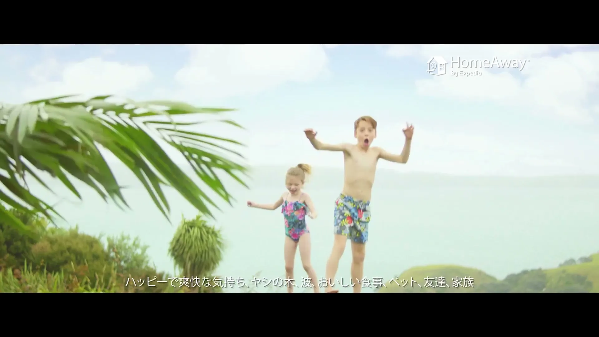 A young boy and a little girl in swimsuits jump excitedly on a grassy cliff overlooking a bright blue ocean, with palm fronds in the foreground.