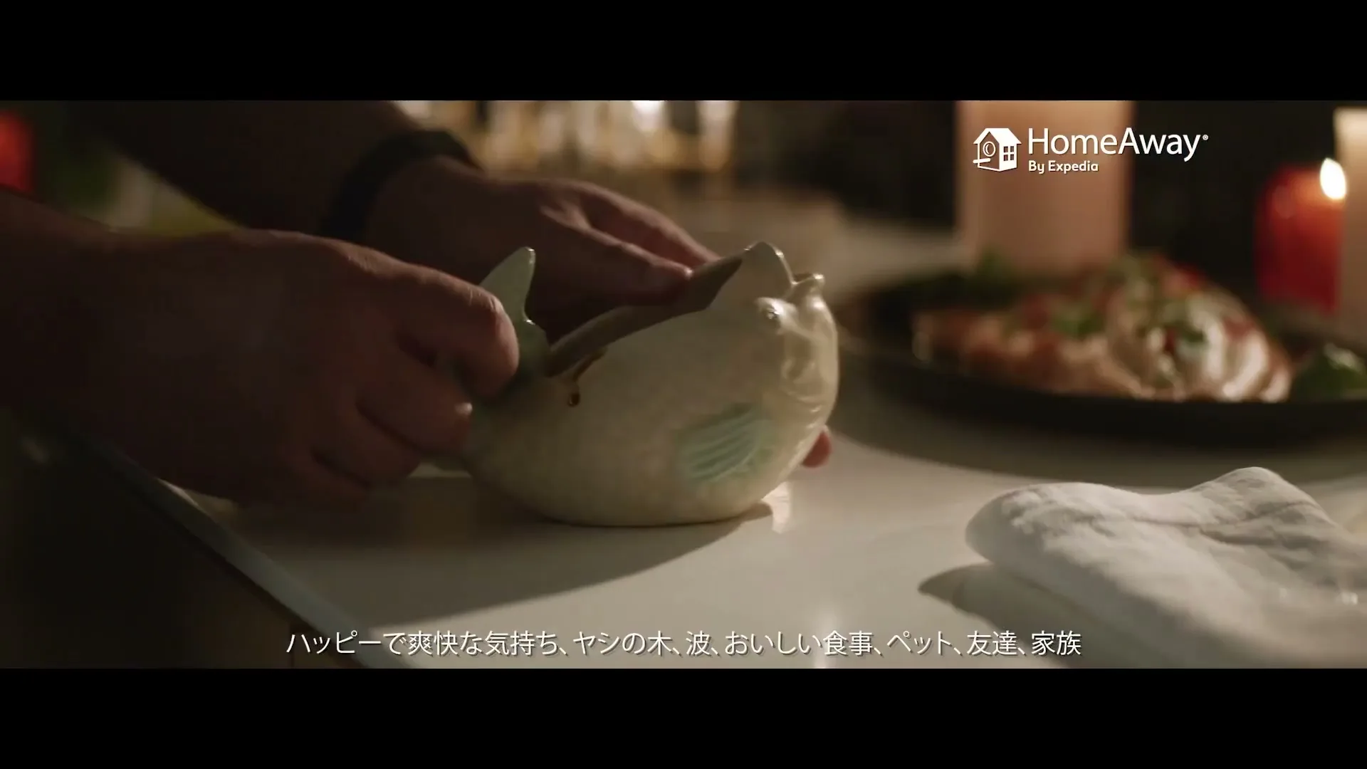 A person's hands are seen placing a fish-shaped dish on a white table, with blurred food and candles in the background, creating a cozy dinner scene.