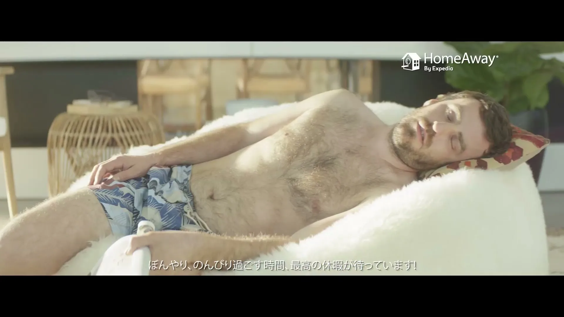 A man with a beard and swim trunks is sleeping on a white beanbag chair in a bright, modern living room.