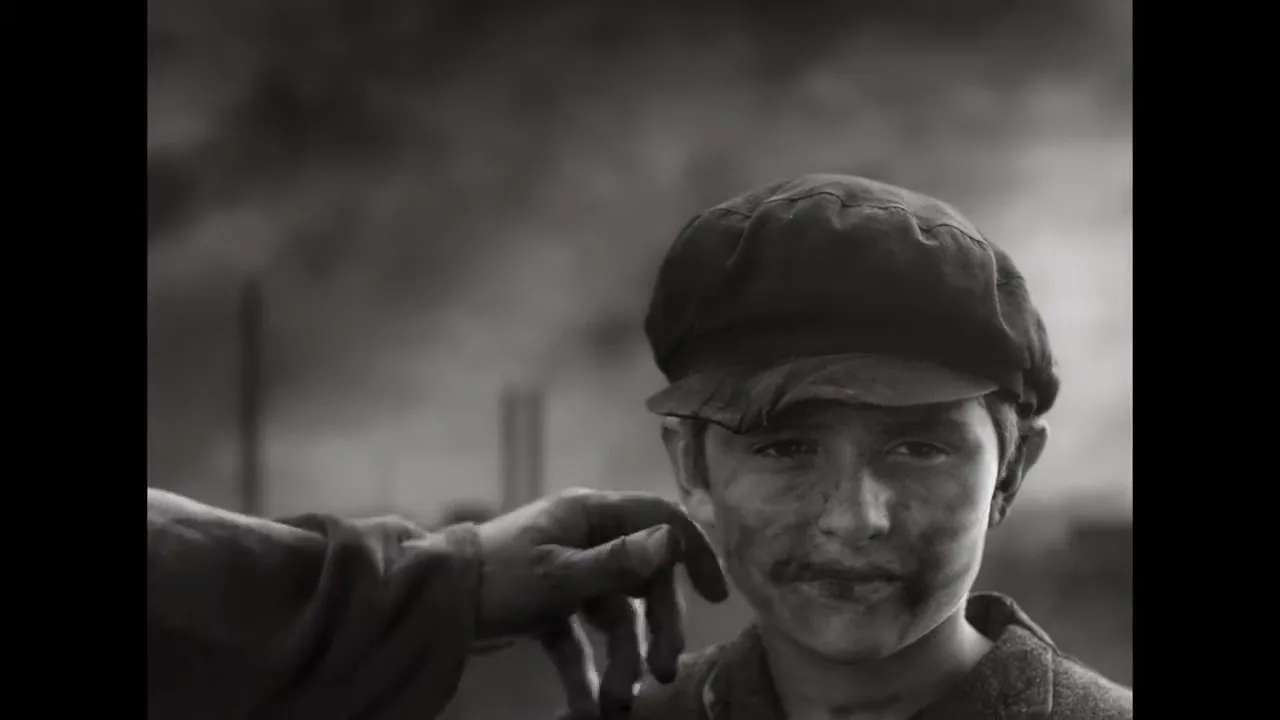 A young boy with a dirty face and cap looks at the camera, then a hand reaches in and pulls his ear, causing him to wince, in a black and white shot.
