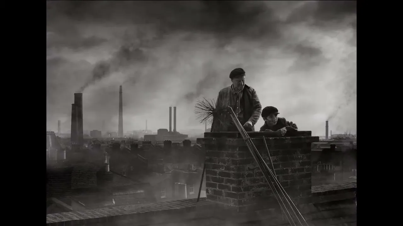 A man and a boy, dressed in old-fashioned clothes, are on a smoky rooftop with industrial chimneys in the background, in a black and white shot.