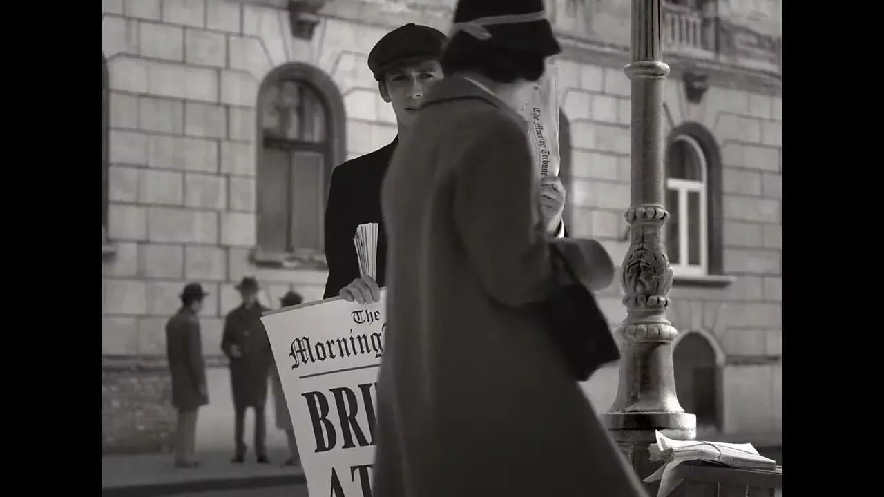 A young male newspaper seller in a cap and suit holds up a newspaper with a headline about war, as a woman in a coat and hat walks past him on a city street.