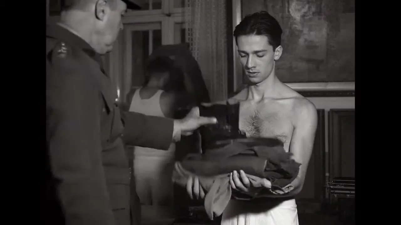 A young man, shirtless and wearing a towel, holds a pile of clothes as an officer hands him a pair of boots in a room.