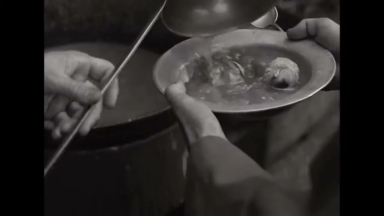 A person's hands hold a metal plate as another person ladles a thin, watery soup with two small lumps into it, in a black and white, close-up shot.