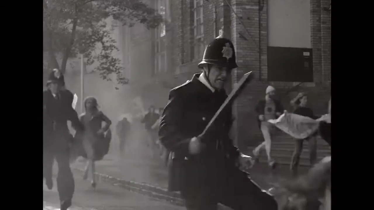 A black and white shot shows a police officer in a helmet and uniform running with a baton, chasing protesters holding a "PEACE" sign on a street.