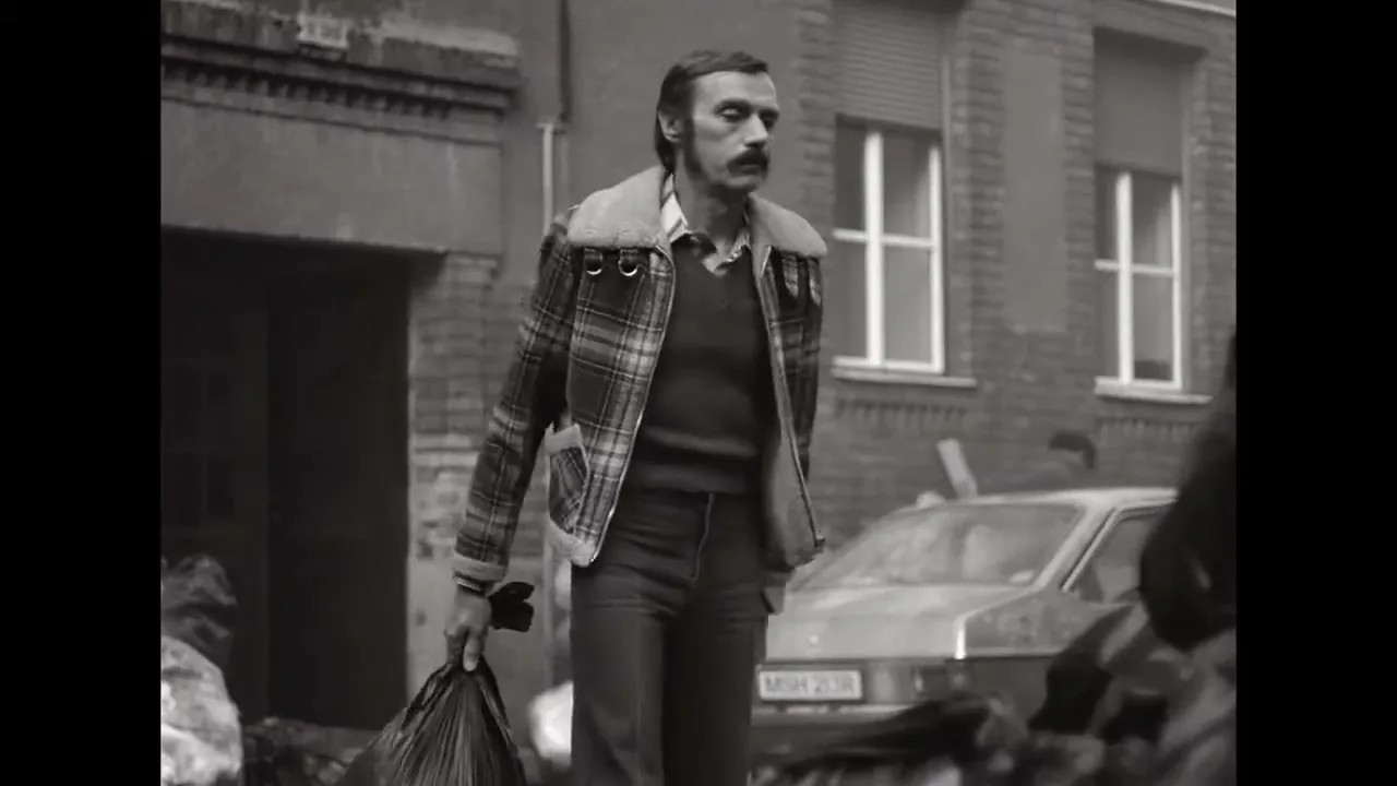 A man with a mustache and plaid jacket walks through a dirty street, carrying a trash bag and looking frustrated in black and white.