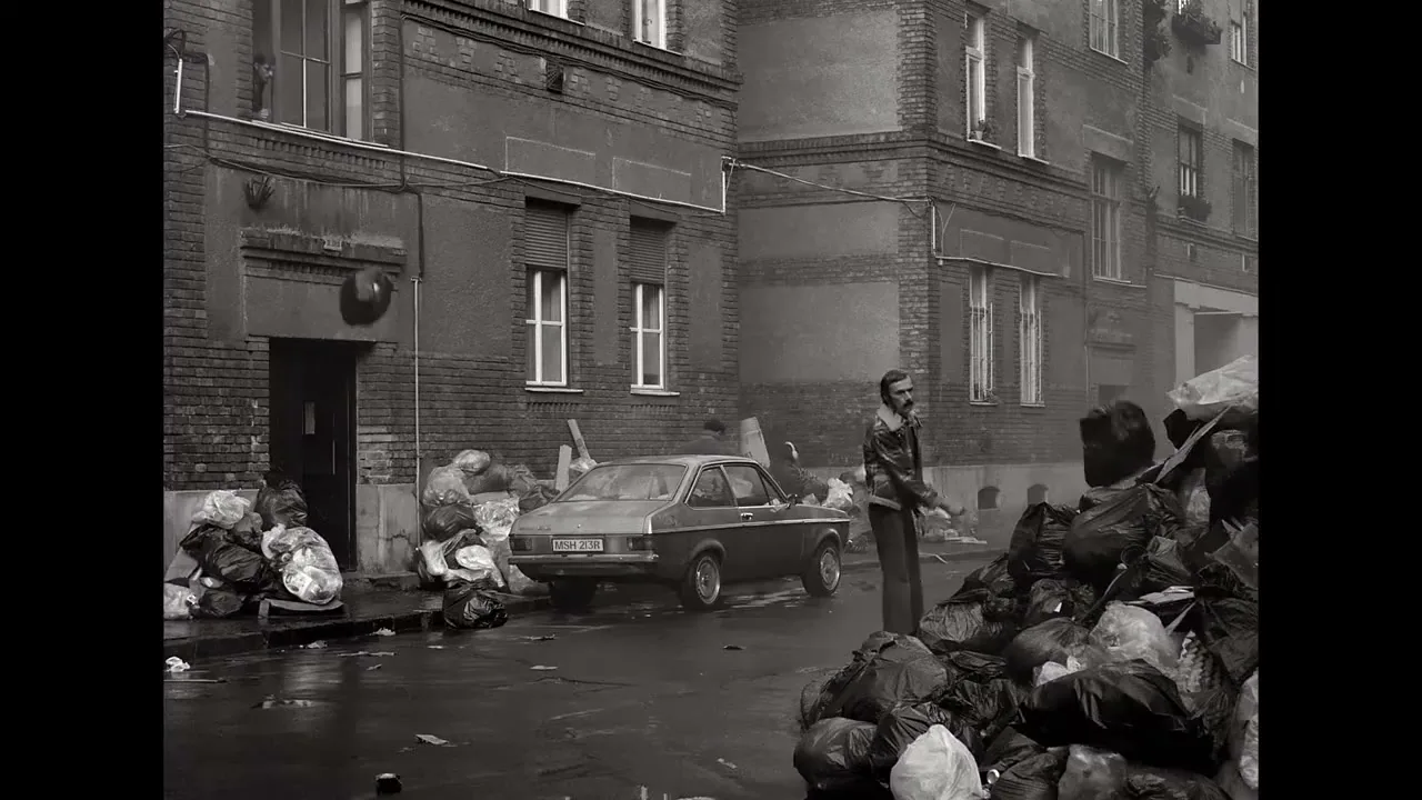 A man in a jacket and trousers stands next to a car on a trash-filled street in a black and white shot.