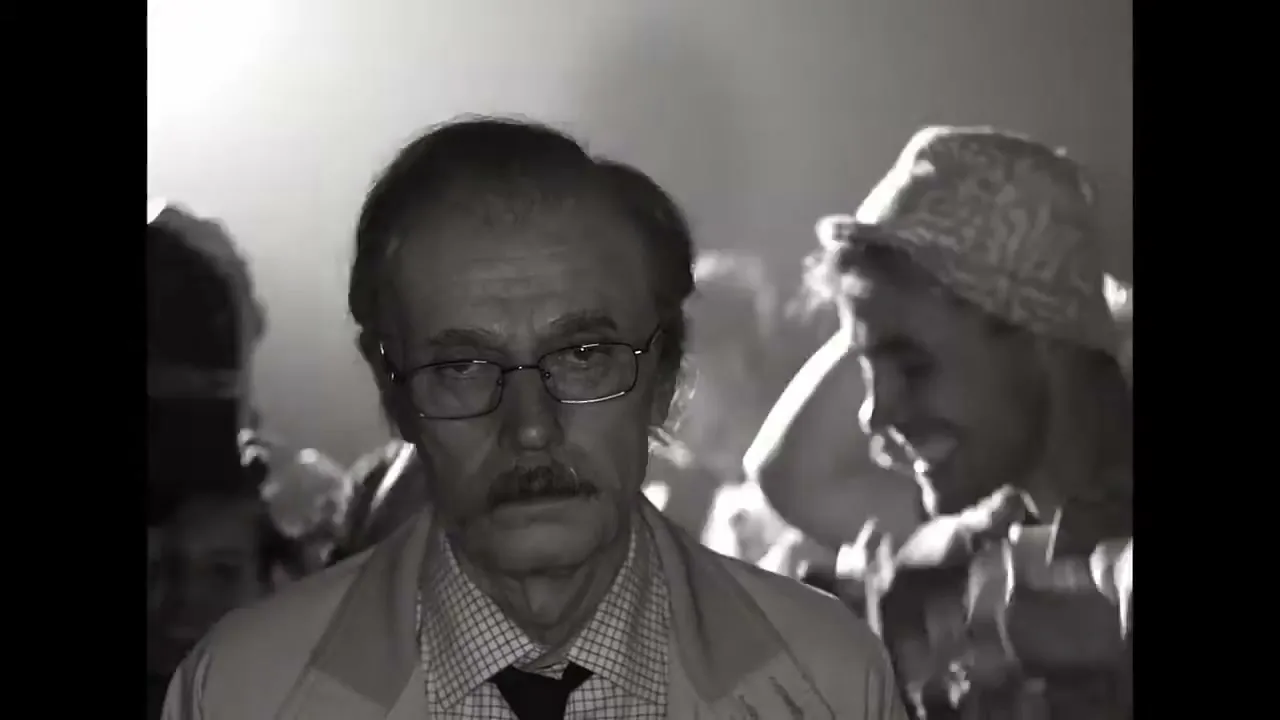 A man with glasses and a mustache looks directly at the camera, while a younger man in a hat laughs in the background at a party.