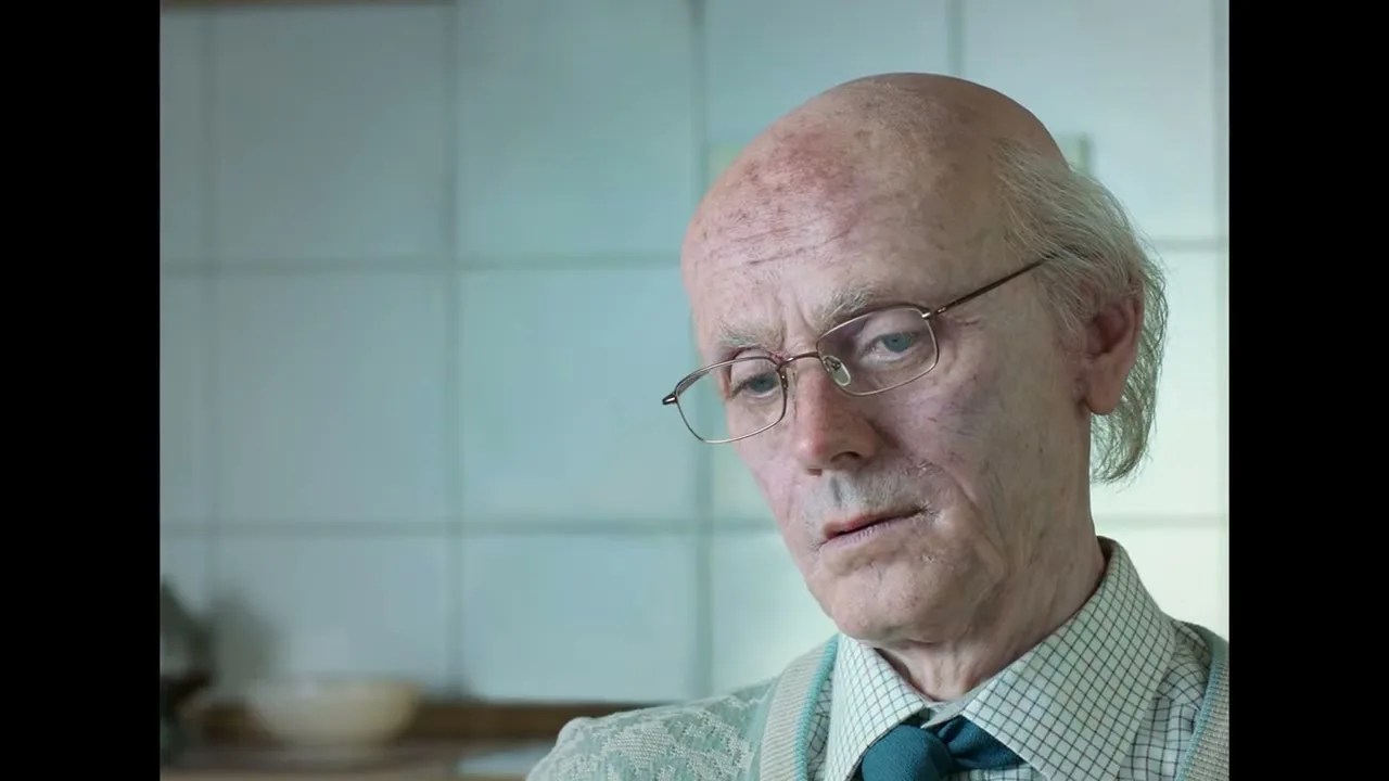 An elderly man with glasses and a receding hairline looks down, then slowly looks up and smiles in a kitchen.