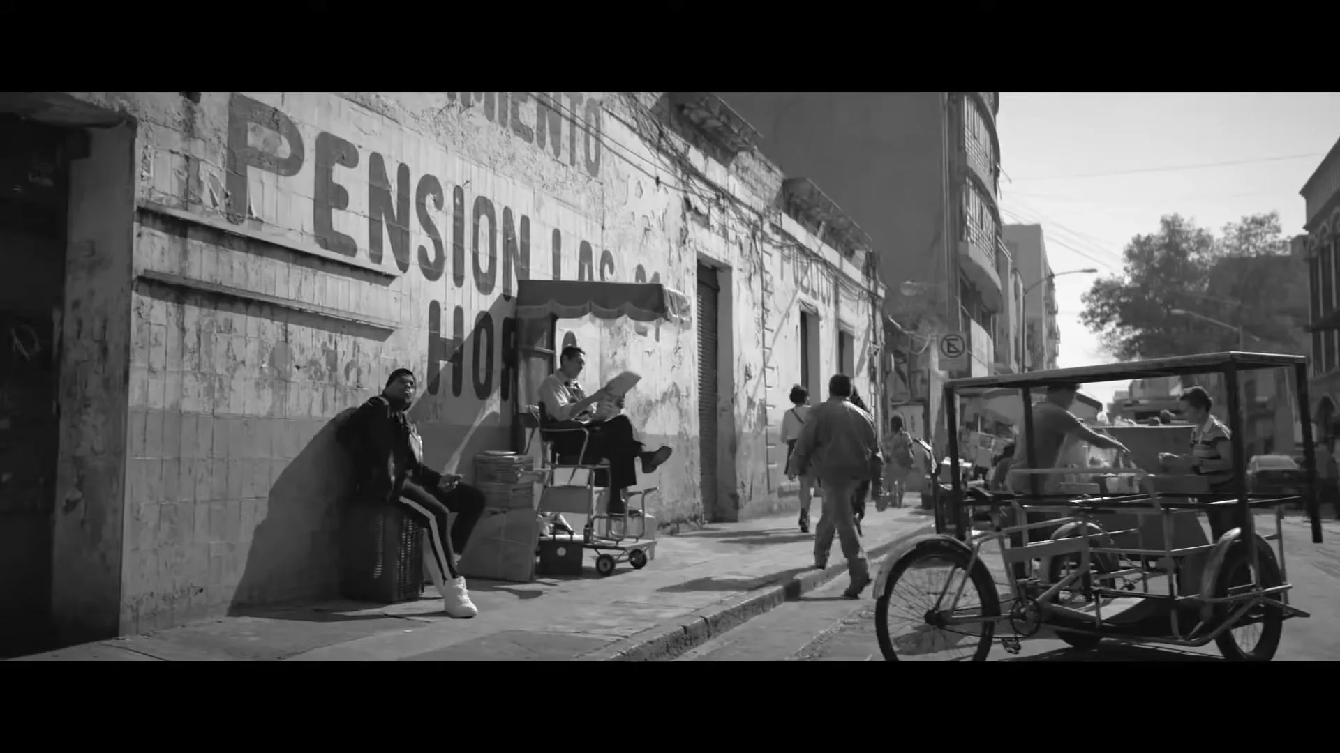 A man in a tracksuit sits against a wall on a busy street with other people walking and a vendor cart in a black and white shot.