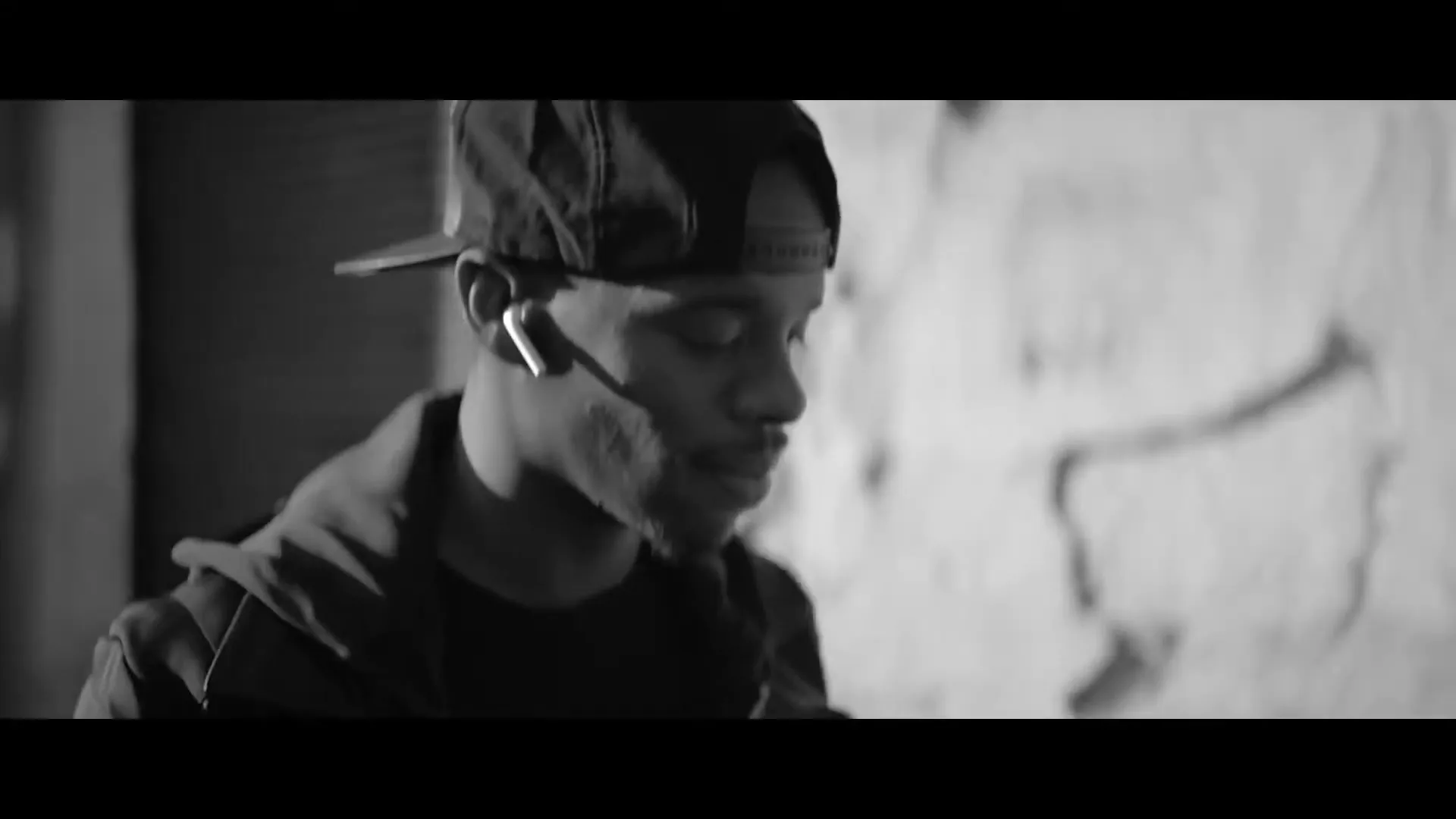 A young man in a baseball cap and hoodie, wearing white earbuds, walks down a city street, looking around with a thoughtful expression.
