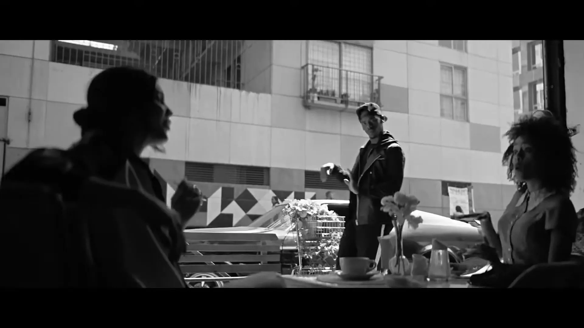 A man in a cap and jacket dances in front of a cafe, while two women sit at a table inside, in a black and white shot.