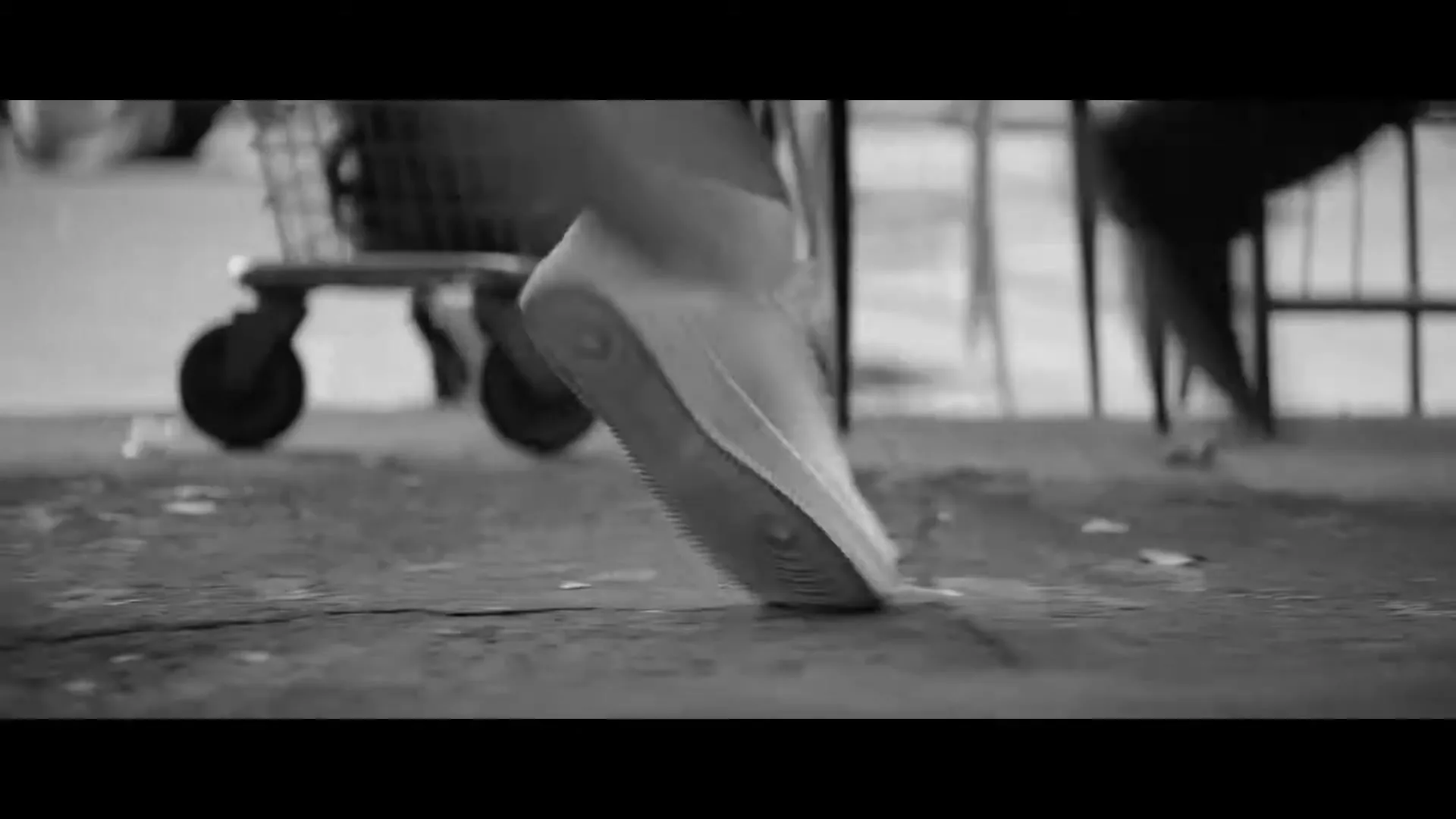 A person's feet in sneakers walk past a shopping cart on a concrete surface in a black and white shot.