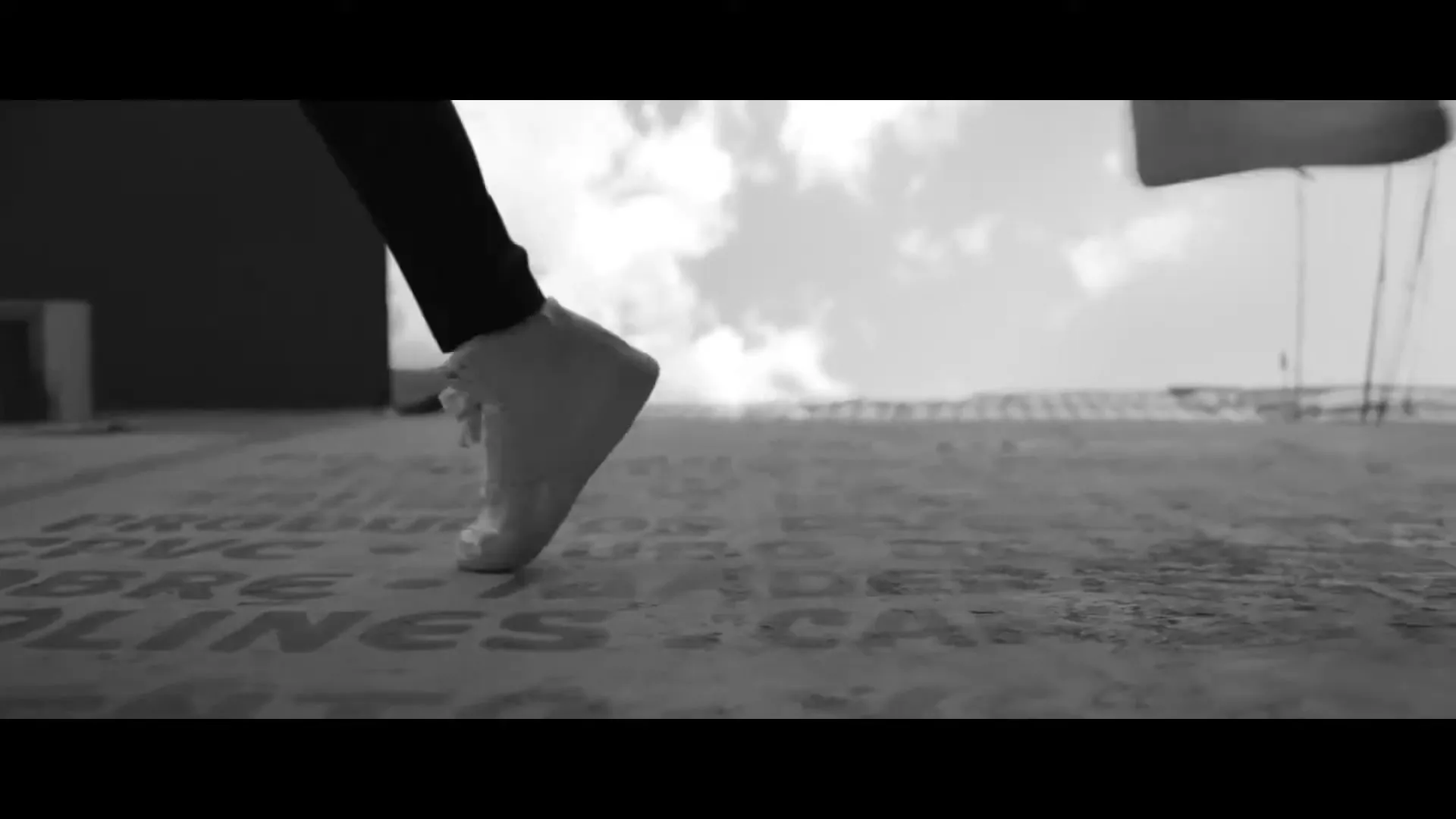 A person's legs and feet in white sneakers dance on a concrete surface with a black baseball cap lying on the ground, under a cloudy sky.