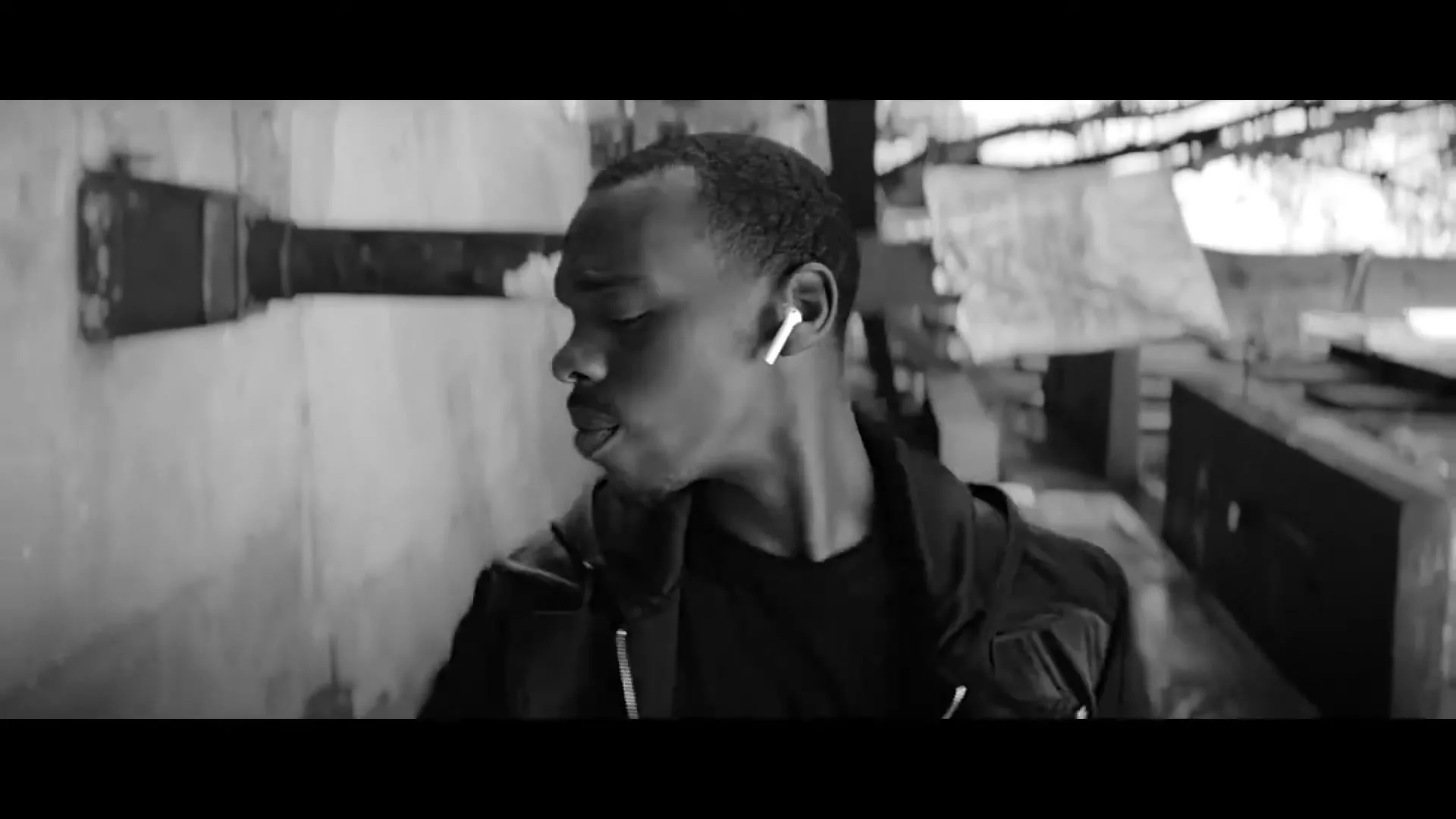 A young Black man in a black hoodie and white AirPods walks through a gritty, industrial-looking alleyway, looking around.