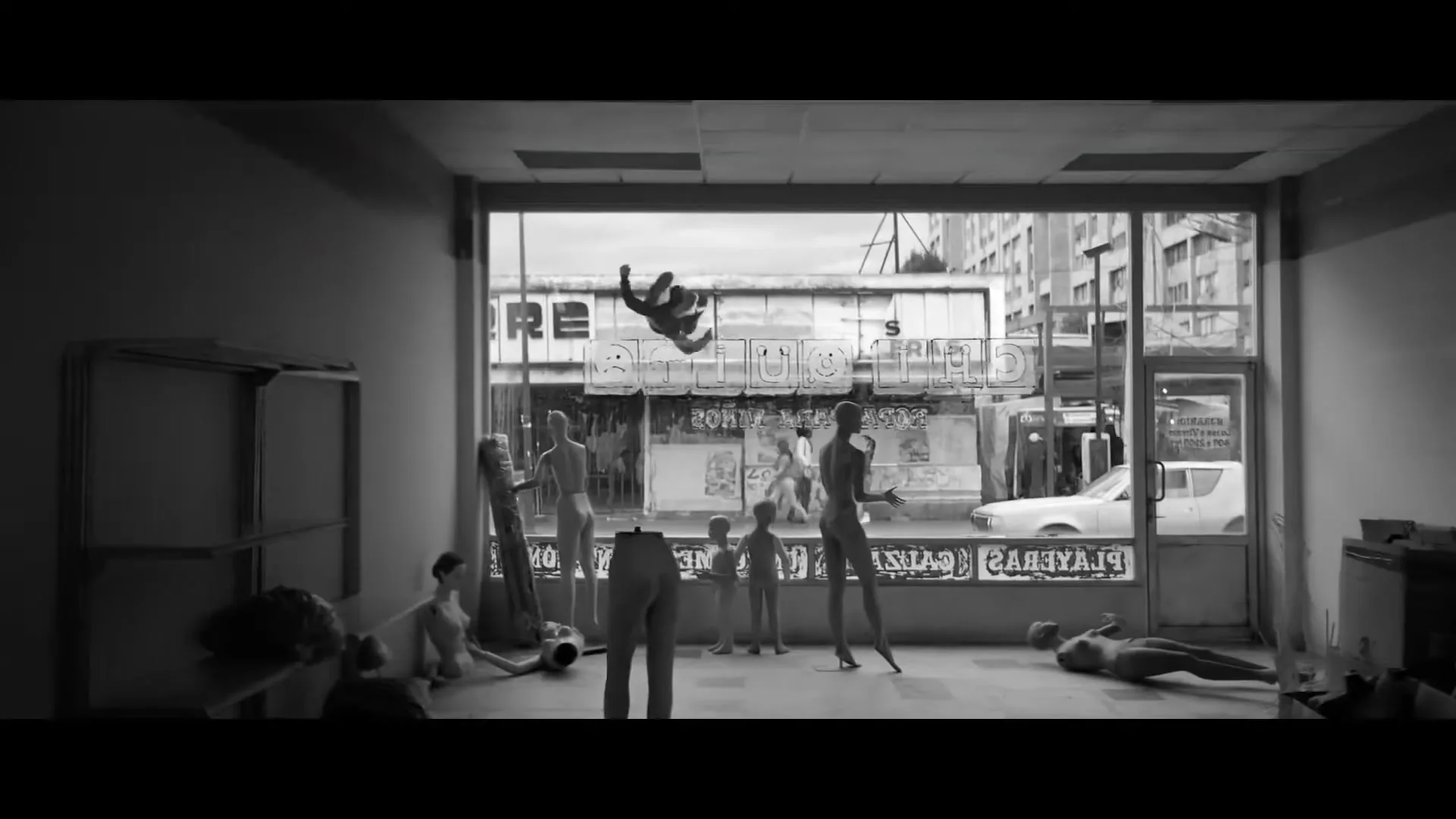 A man in a dark suit jumps and spins in the air outside a storefront window, surrounded by mannequins inside, in a black and white shot.