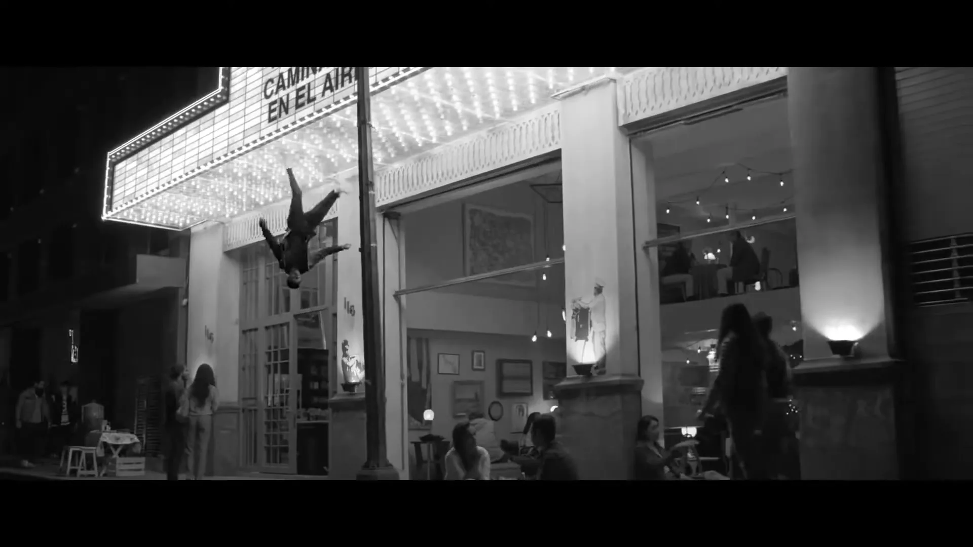 A man in a dark suit hangs upside down from a lamppost in front of a brightly lit theater marquee and a cafe at night.