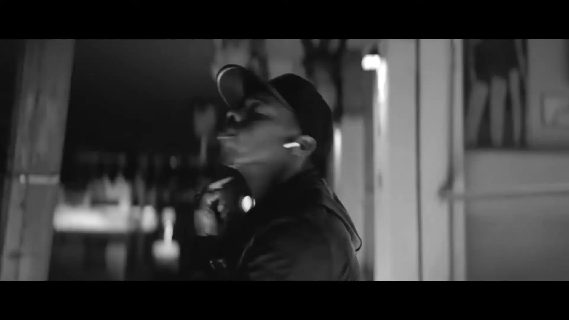 A young Black man in a baseball cap and jacket, wearing AirPods, walks and looks around in a dark, urban exterior at night.