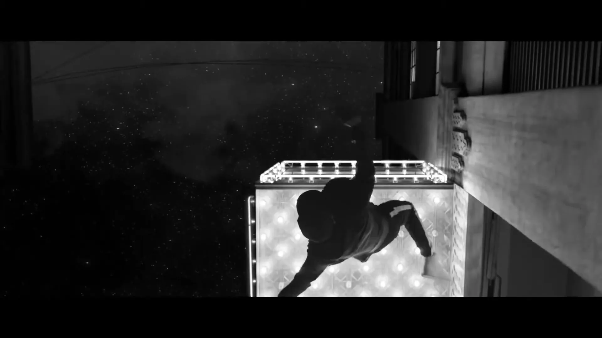 A man in a hoodie and sweatpants is seen from an overhead angle, falling through the air next to a building at night.