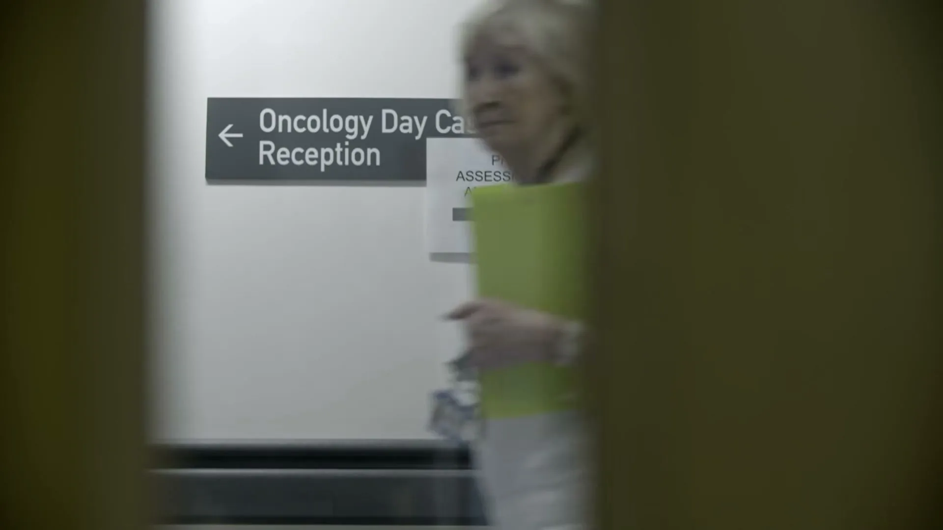 A blurred shot of a hospital corridor with a sign for "Oncology Day Case Unit Reception" and "Pre-Assessments and Clinics" as a woman walks past.