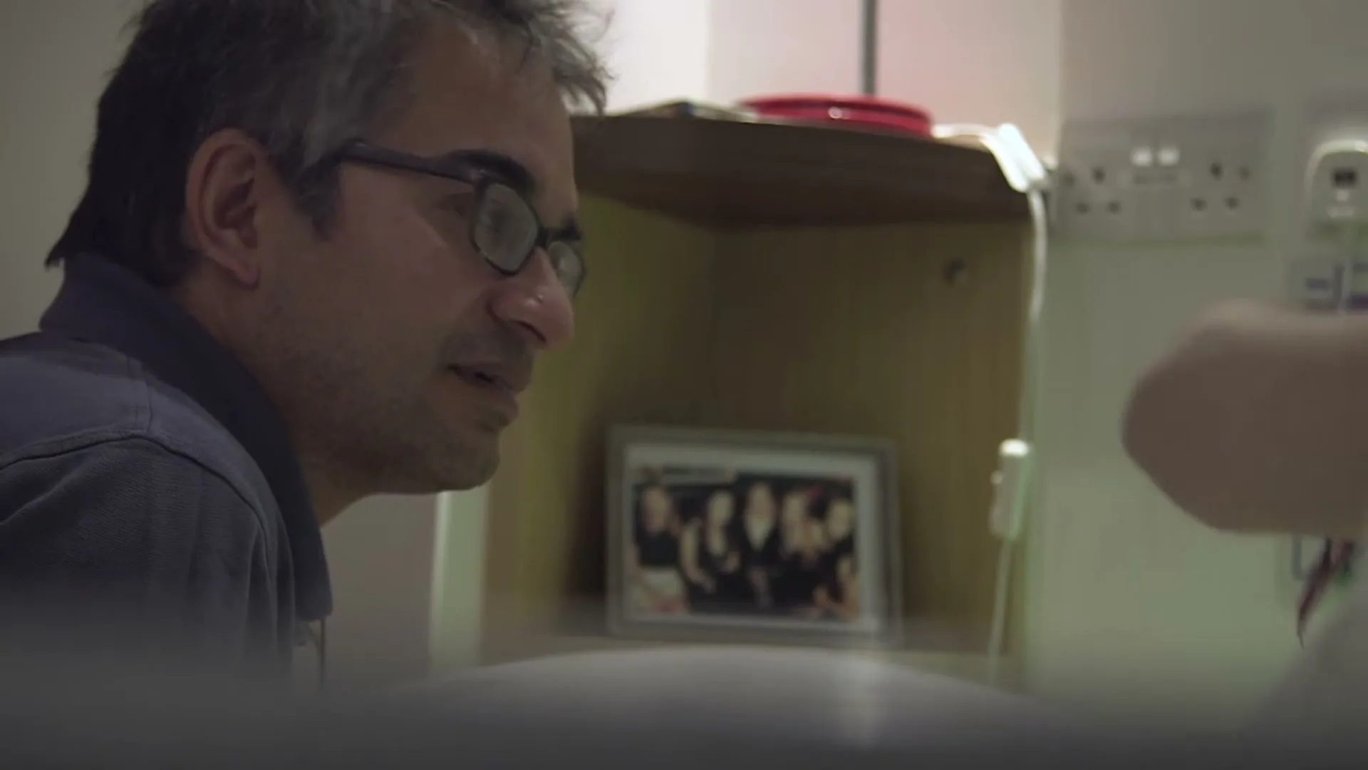 A man with glasses and grey hair looks at a framed photo of a group of people in a hospital room, pointing at it.