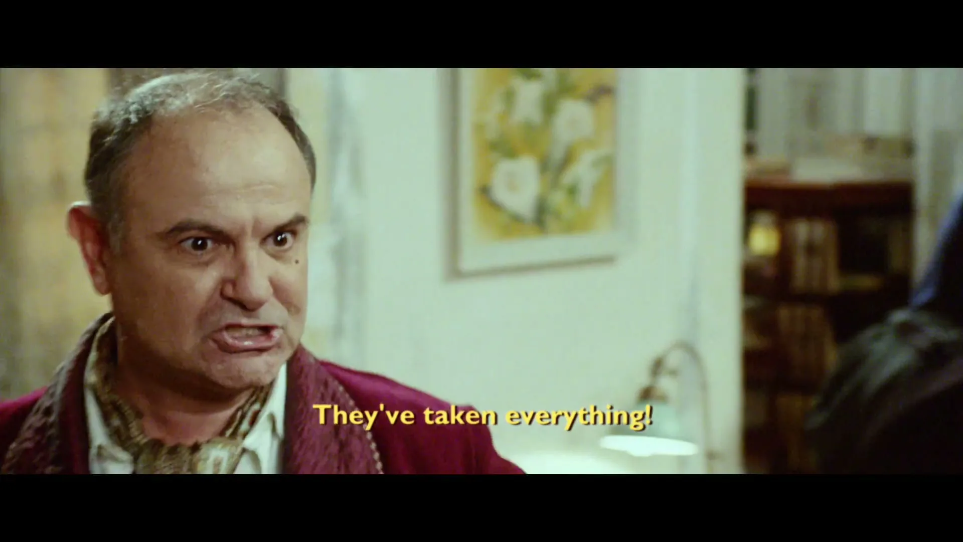 A man in a red robe with a shocked expression yells at someone off-screen in a dimly lit room, while another man stands in the background.