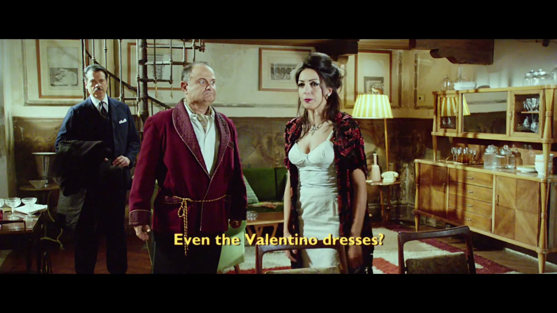 A man in a robe and a woman in a dress argue in a living room, while another man in a suit stands in the background.