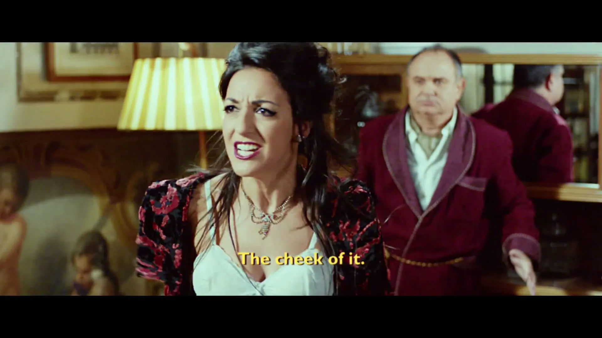 A woman in a white bra and a red and black top argues with a man in a red robe in a living room, both looking angry.