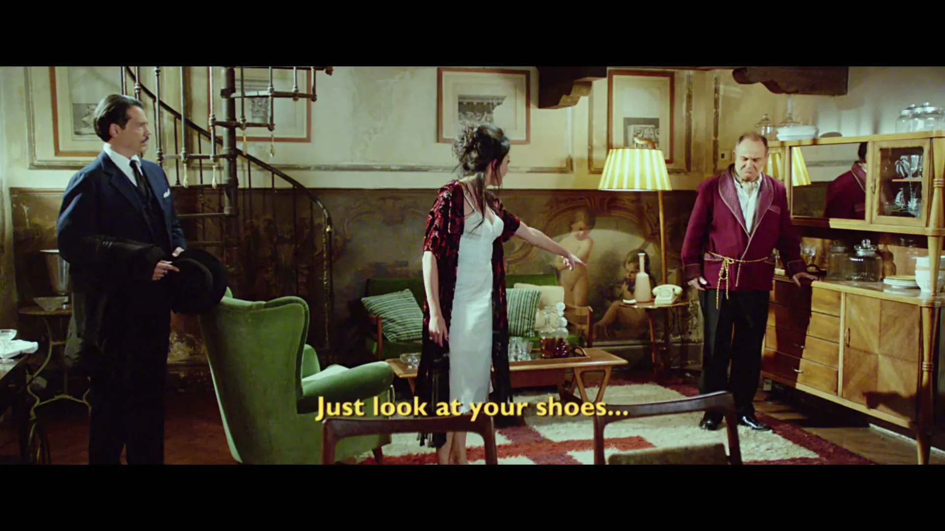 A woman in a white dress points at a man in a red robe, while another man in a suit watches in a vintage Italian living room.