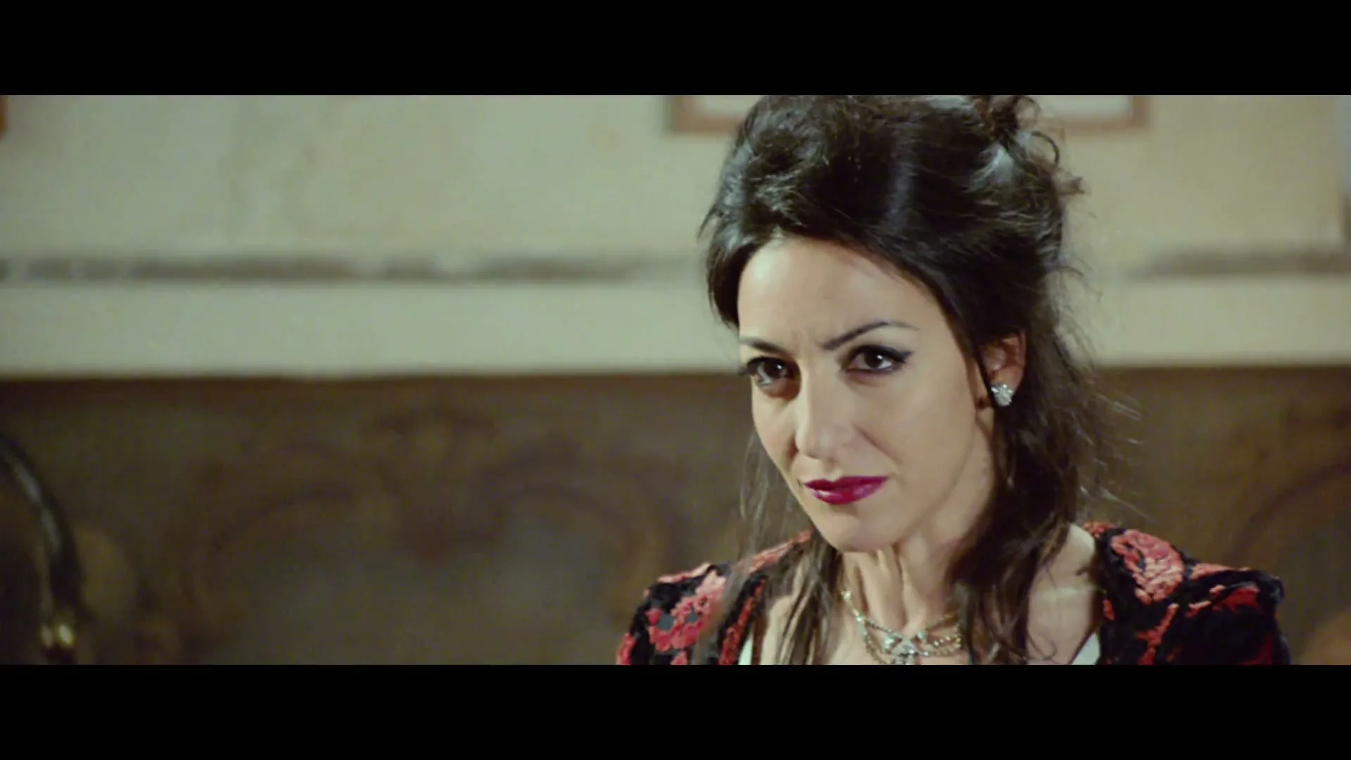 A woman with dark hair and a patterned top looks around nervously in an ornate room, then looks at the camera and asks "Him?".