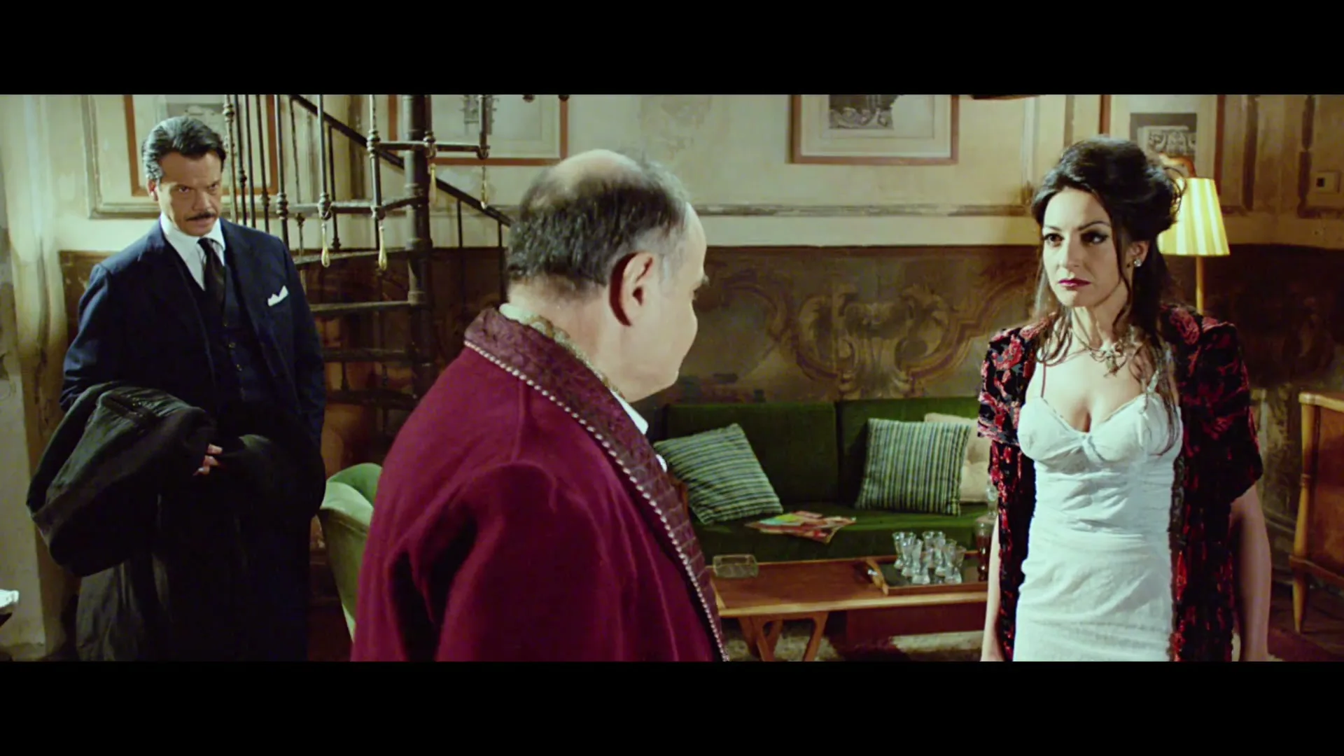 A man in a suit, a man in a robe, and a woman in a white dress and red shawl are in a room with a spiral staircase and a green couch.