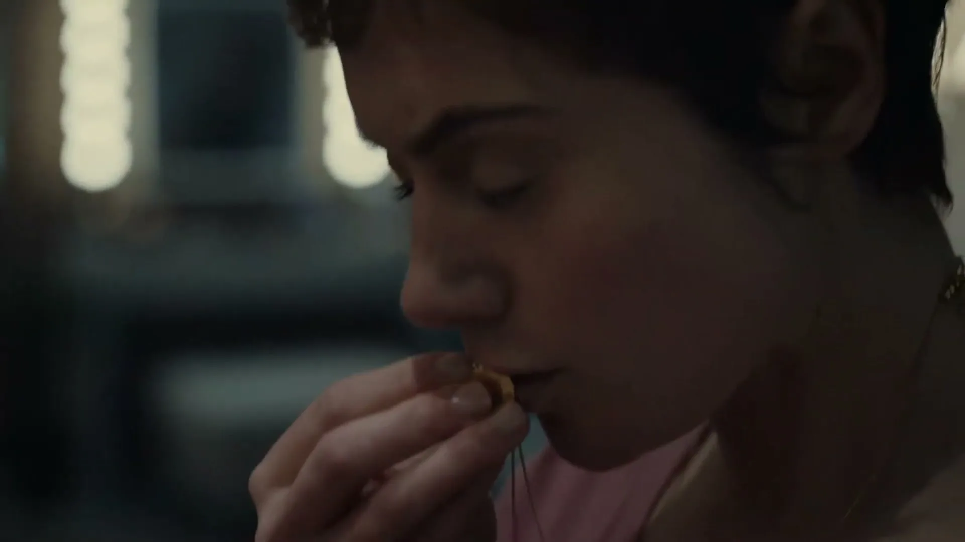 A young woman with short dark hair in a pink top puts on a small gold necklace in a dimly lit room.