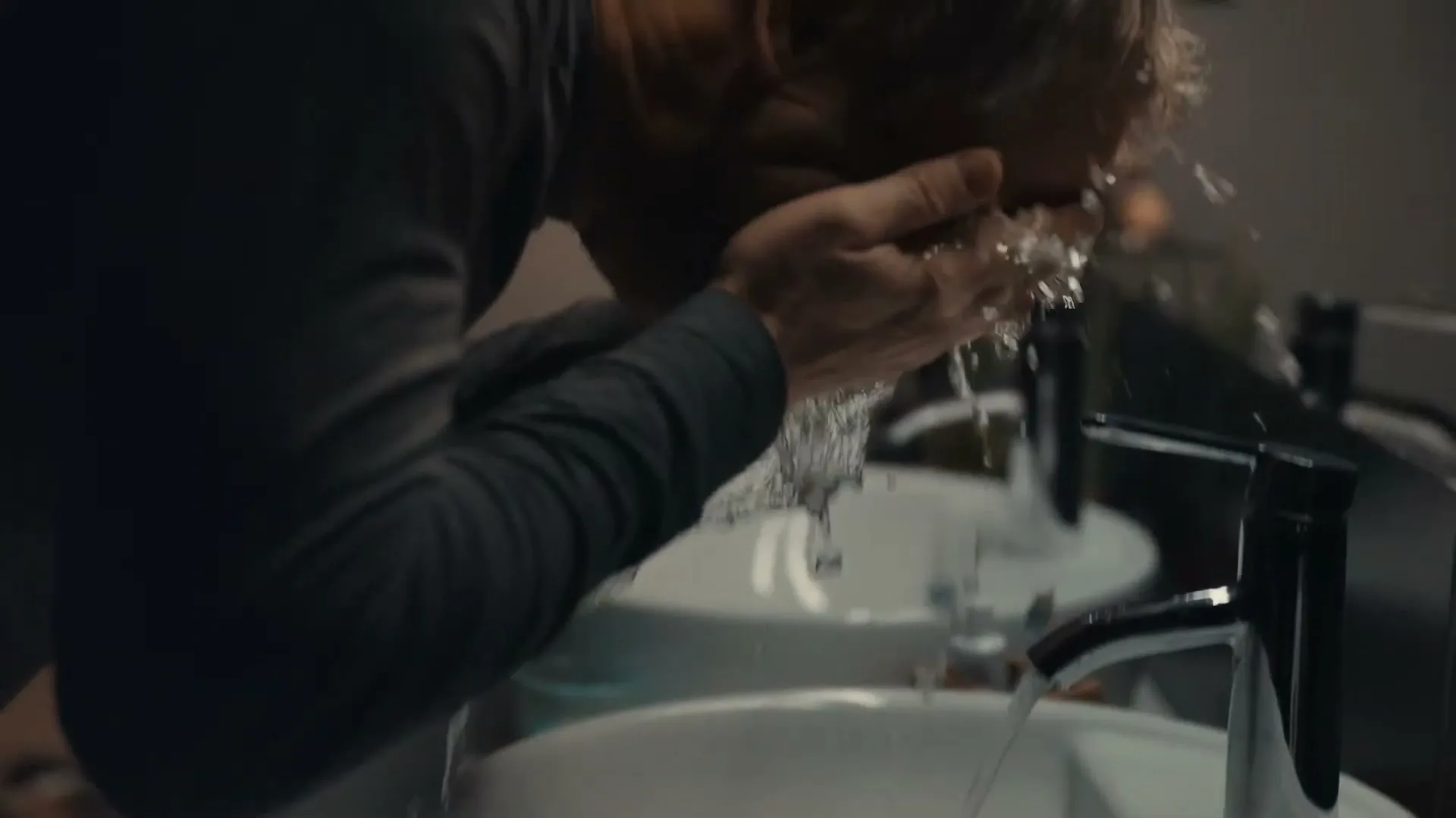 A man with short blonde hair washes his face in a white sink with running water in a modern bathroom.
