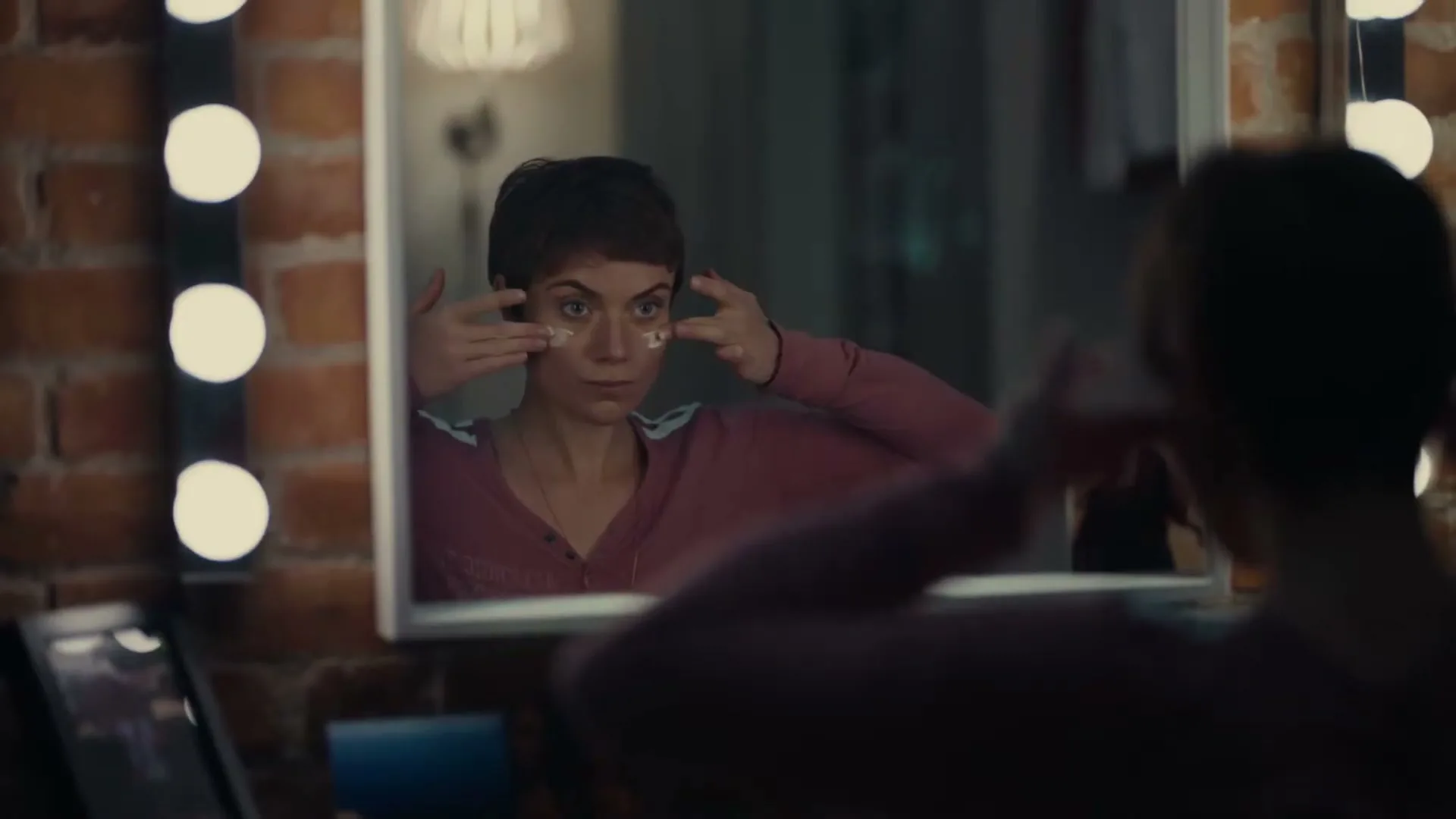 A woman with short hair applies eye cream in a mirror, illuminated by vanity lights, in a bedroom at night.