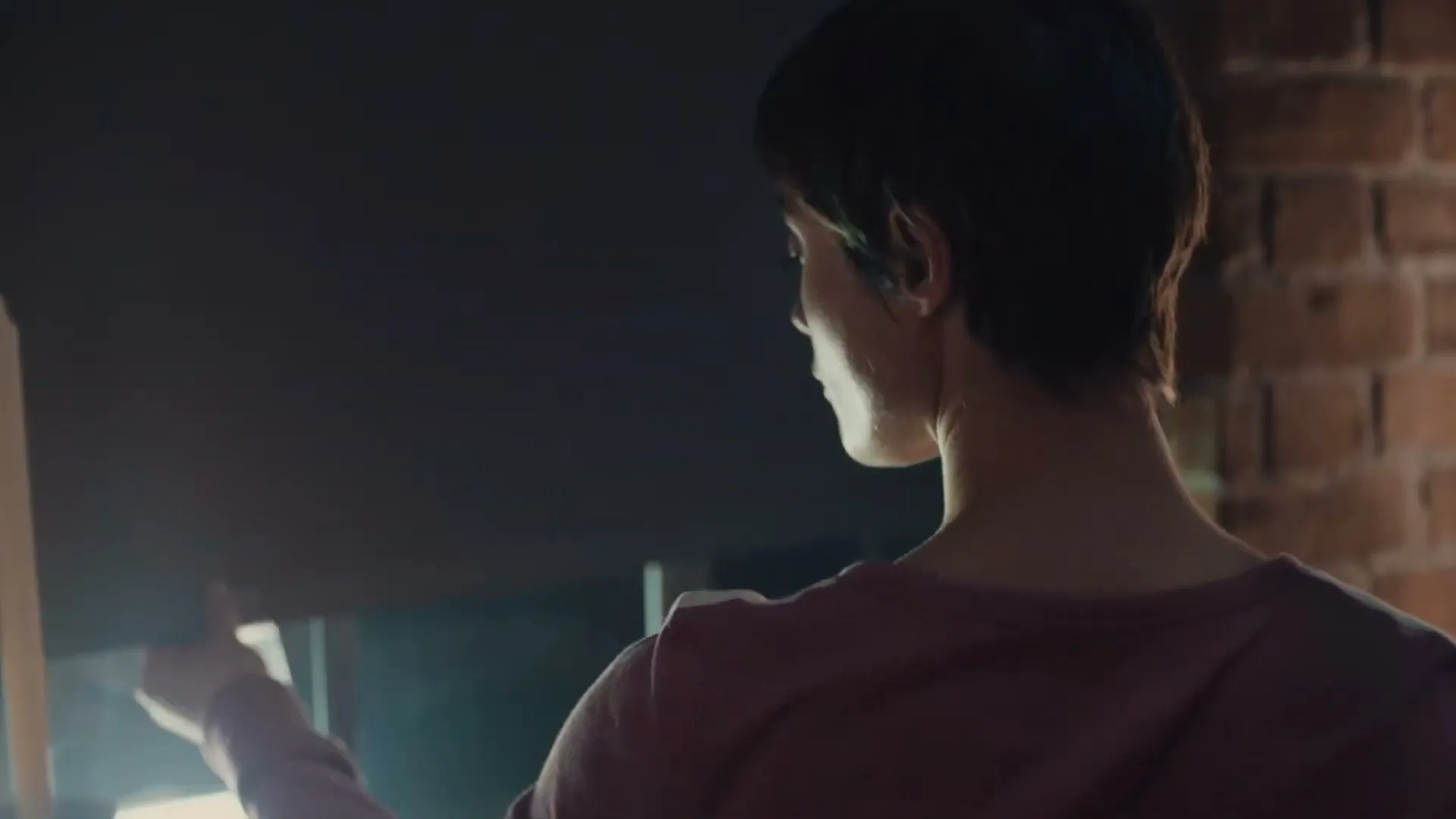 A woman with short dark hair, seen from behind, closes a dark window blind in a room with a brick wall.