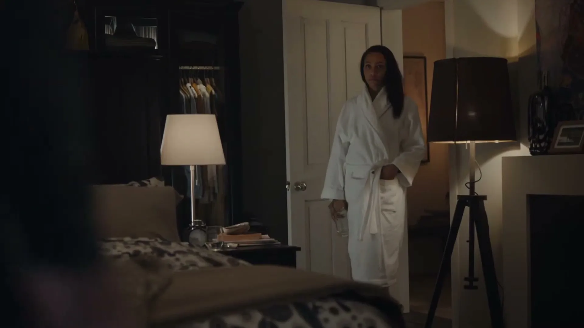 A woman in a white robe stands in a dimly lit bedroom, holding a glass of water, looking towards the bed.