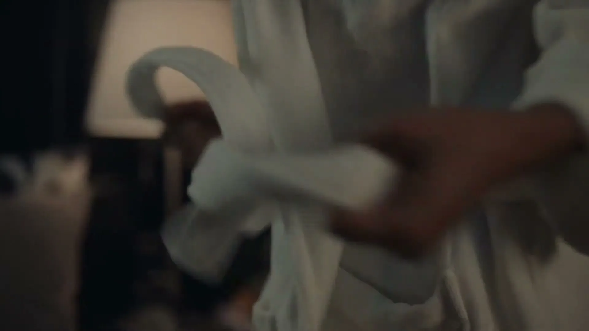 A person's hands are seen tying a white bathrobe in a dimly lit room, with a lamp in the background.