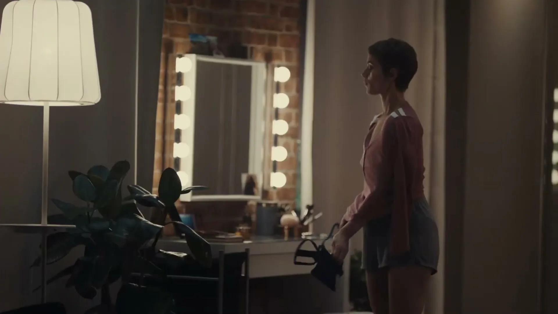 A woman with short hair, wearing a pink top and shorts, walks past a vanity mirror with lights in a bedroom at night.