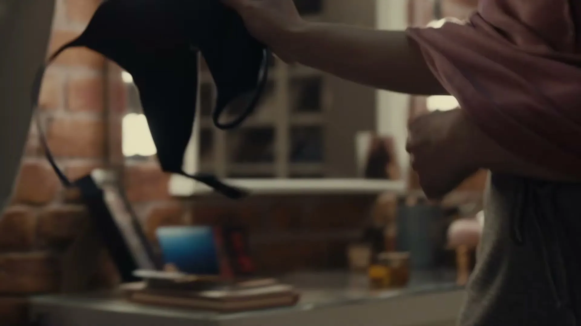 A person in a pink shirt and grey shorts takes off a black bra in front of a vanity mirror with lights in a bedroom.
