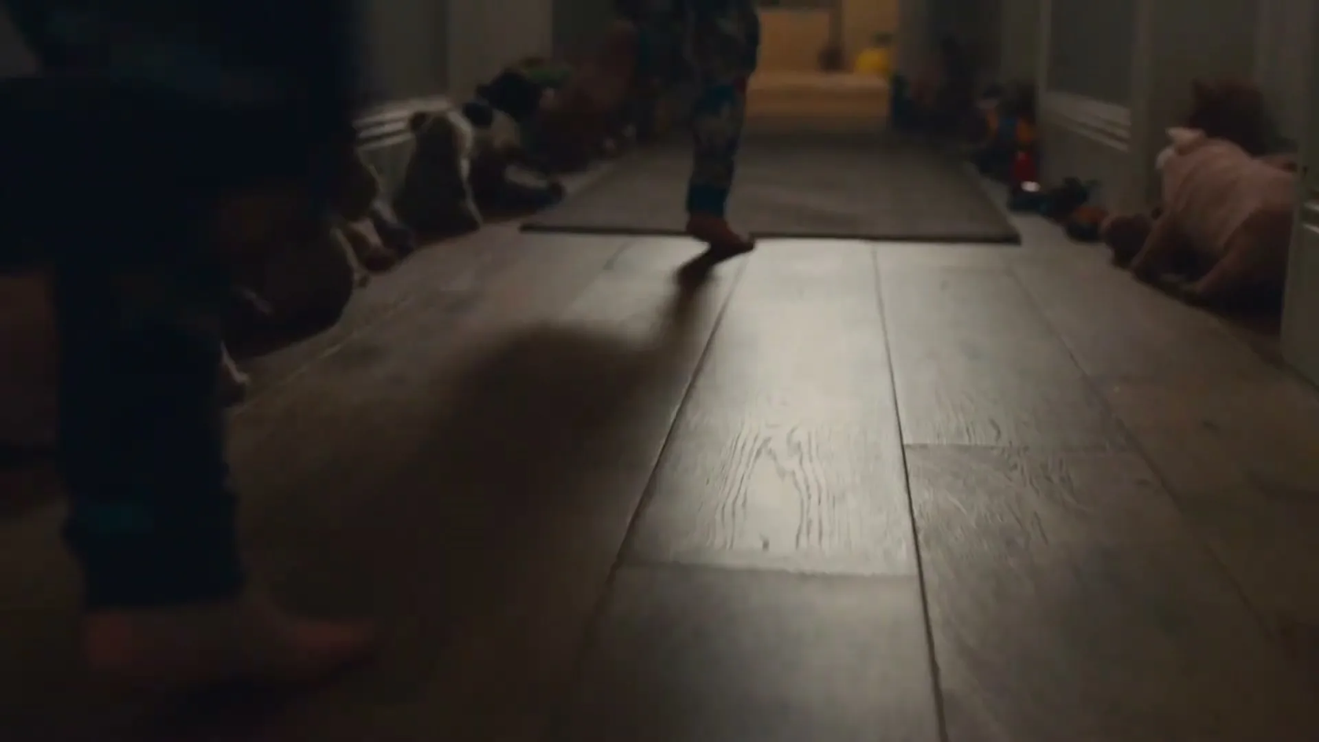 A child in pajamas runs barefoot down a dimly lit hallway, with stuffed animals lining the floor on both sides.